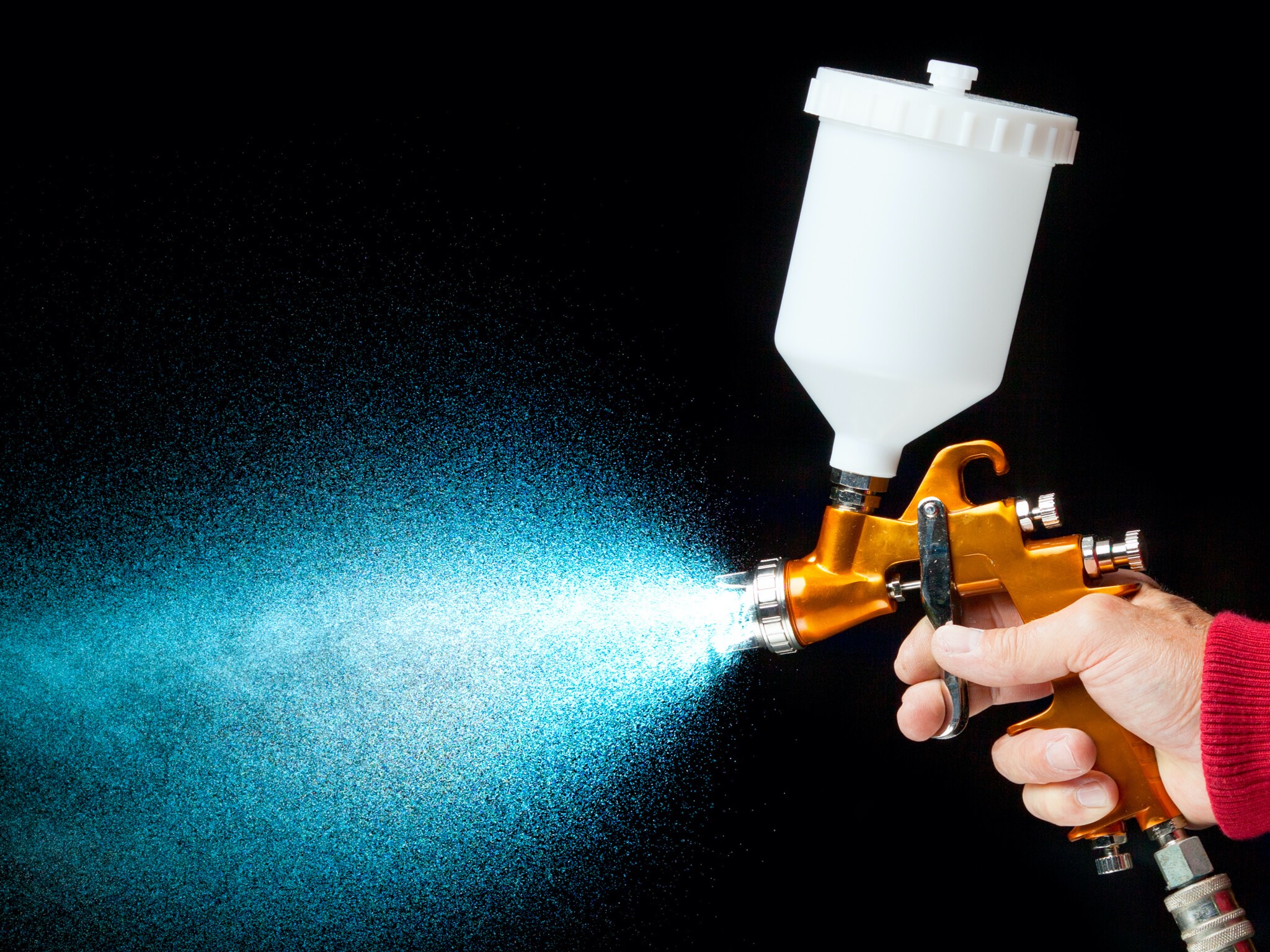 Złoty pistolet natryskowy w dłoni rozpyla błękitną farbę na czarnym tle, widoczny przewód doprowadzający powietrze.