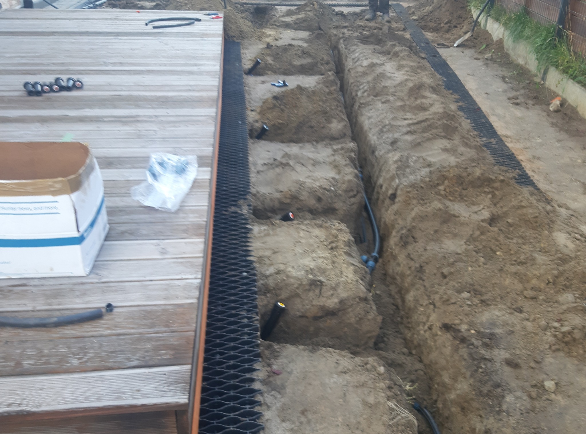 Wykopane rowy pod system nawadniania ogrodu, widoczne rury i złączki, obok drewniany taras z elementami systemu.