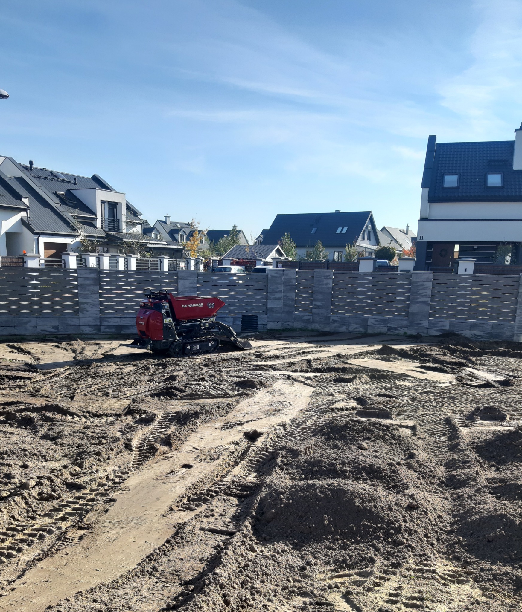 Czerwona minikoparka Yanmar na przygotowanym terenie pod trawnik, widoczne ślady opon w piasku, w tle nowoczesne domy jednorodzinne i ażurowy płot.