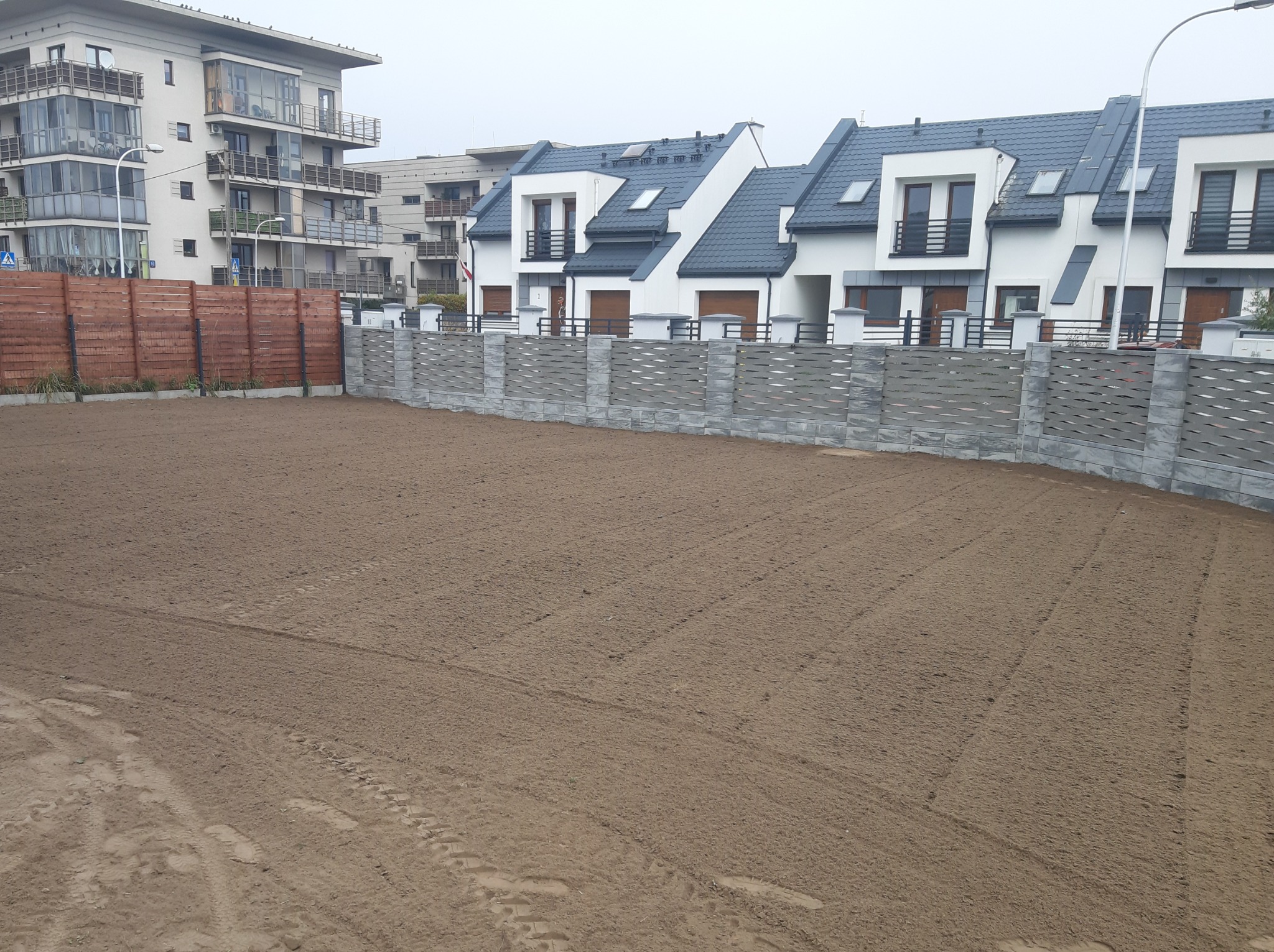 Przygotowany teren pod trawnik z widocznymi śladami po maszynach, w tle nowoczesne domy szeregowe i apartamentowiec.