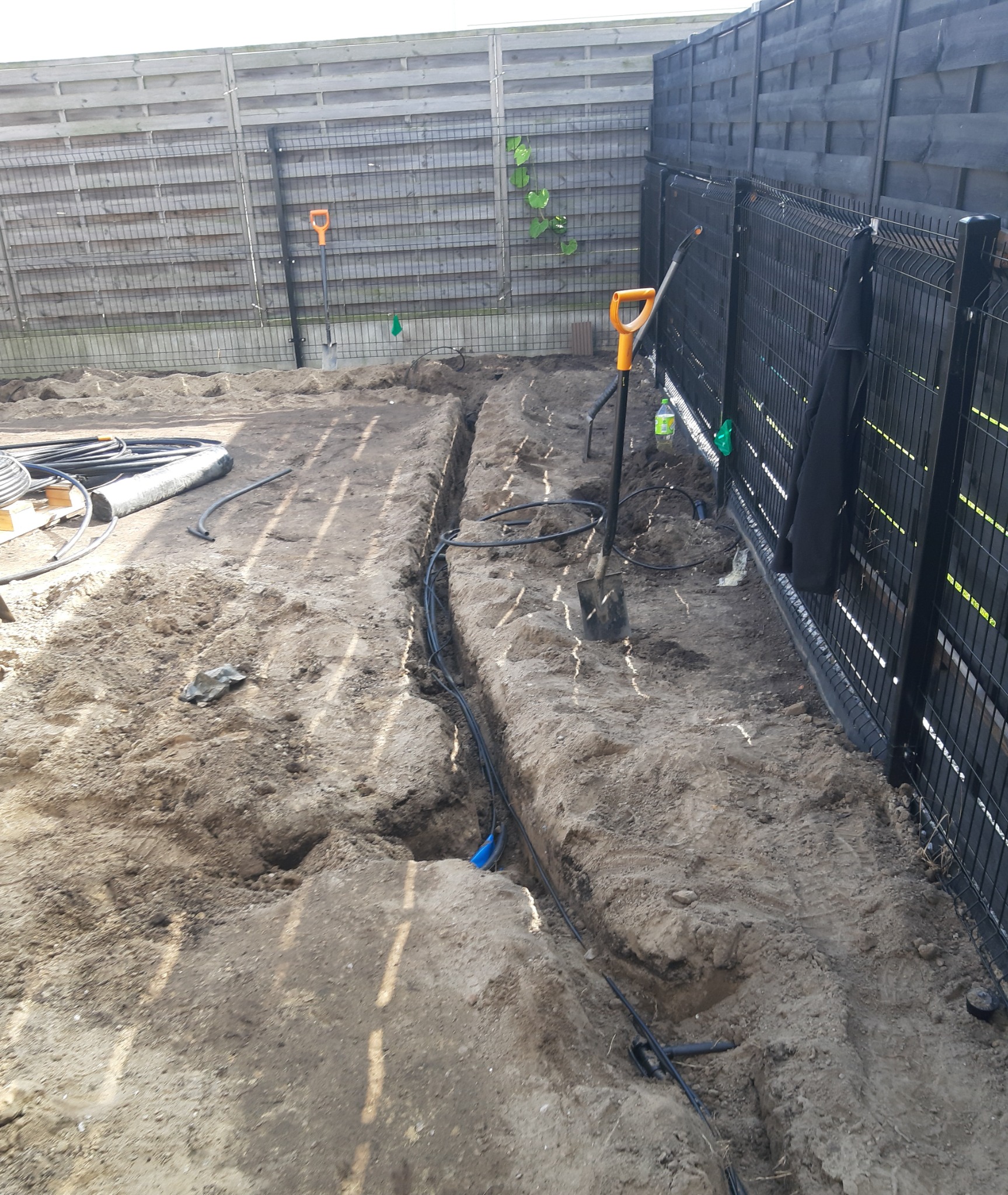 Wykopane rowy w ziemi pod system nawadniania, widoczne rury i dwie łopaty z pomarańczowymi uchwytami, czarne ogrodzenie panelowe w tle.