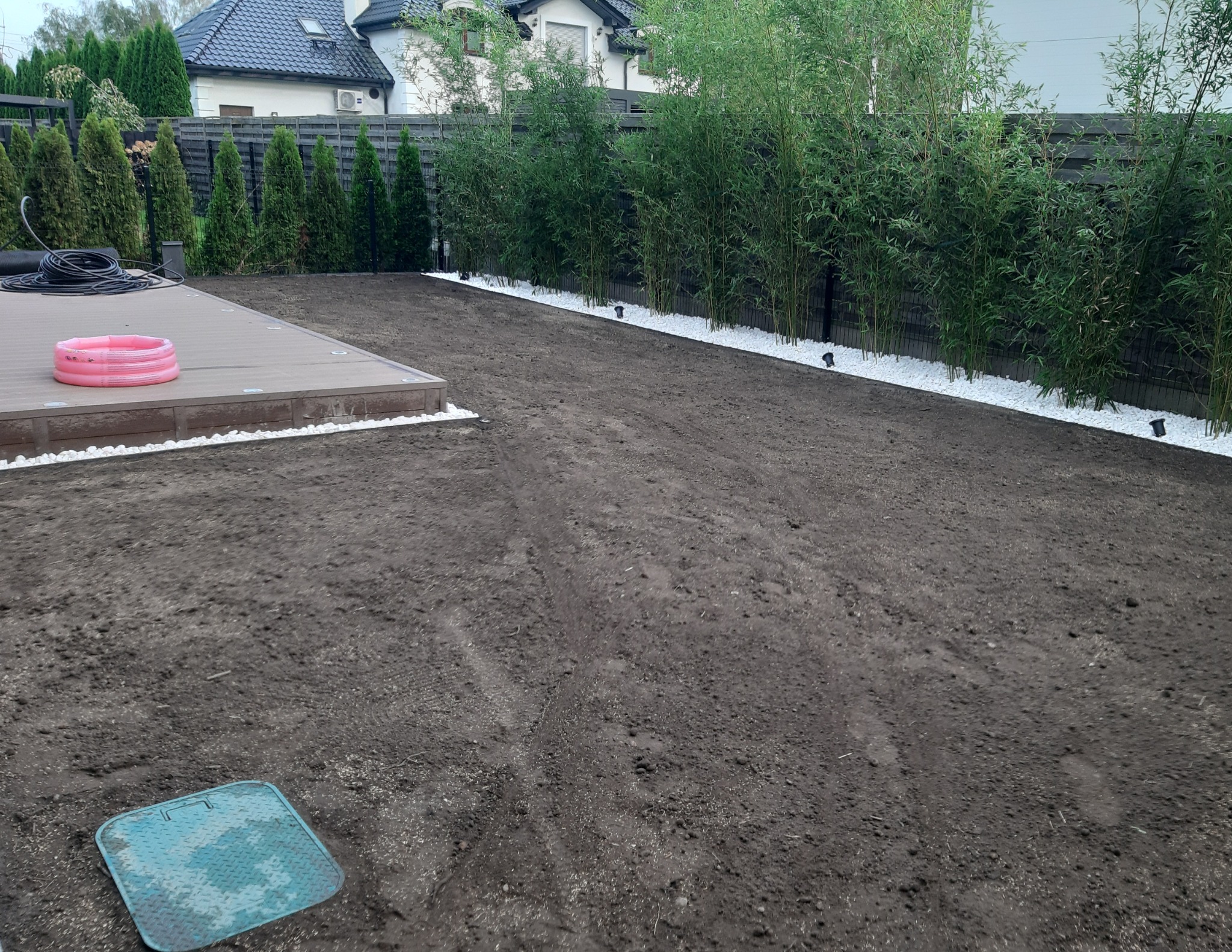 Przygotowany teren pod trawnik z rolki, widoczny system nawadniania i dekoracyjne białe kamienie wzdłuż ogrodzenia, obok tarasu z małym, różowym basenem.