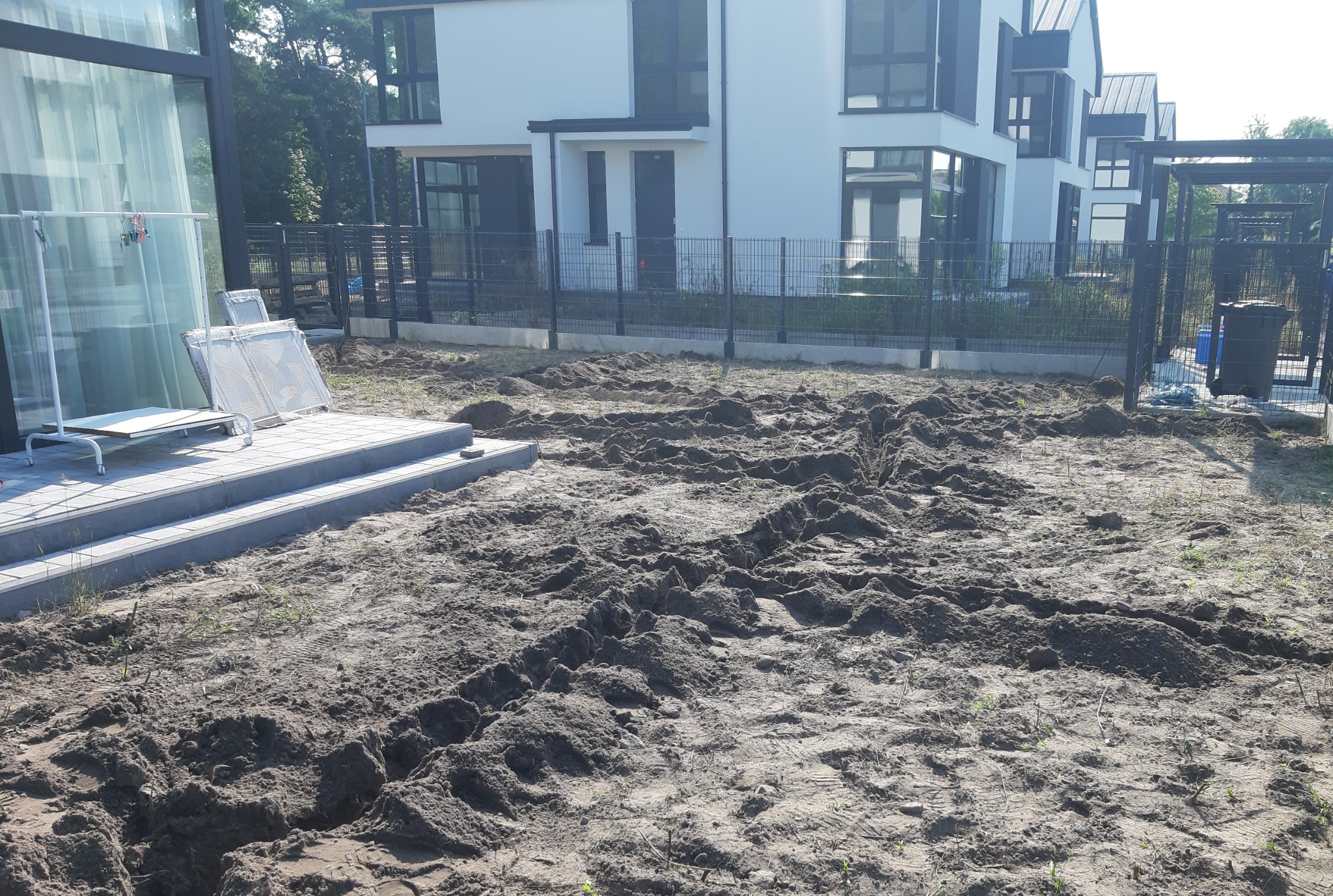 Przygotowany teren pod trawnik, świeżo przekopana ziemia z widocznymi śladami maszyn, w tle nowoczesne domy jednorodzinne i metalowe ogrodzenie.