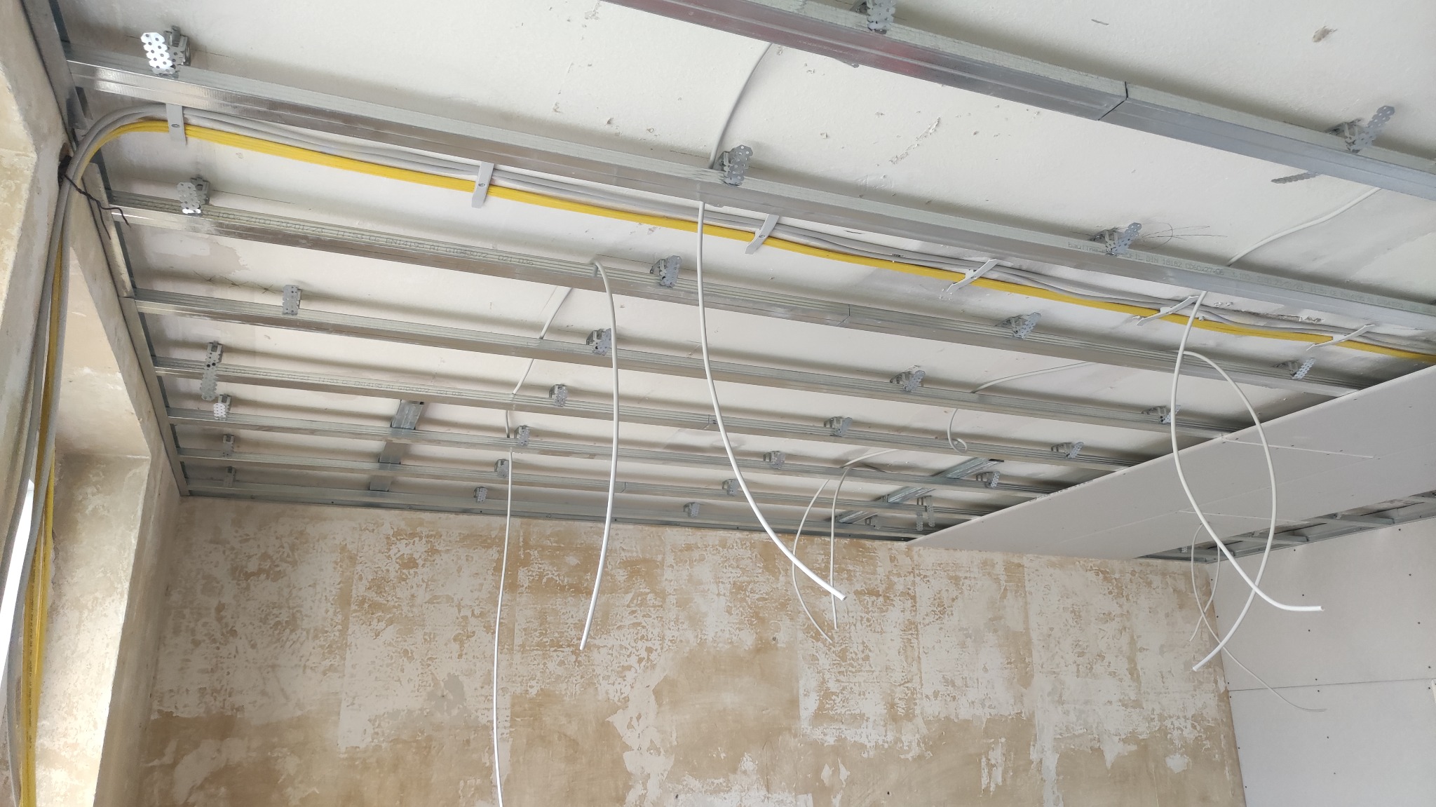 Montaż metalowej konstrukcji sufitu podwieszanego z widocznymi przewodami elektrycznymi i częściowo zamontowaną płytą gipsowo-kartonową w pomieszczeniu z surowymi ścianami.