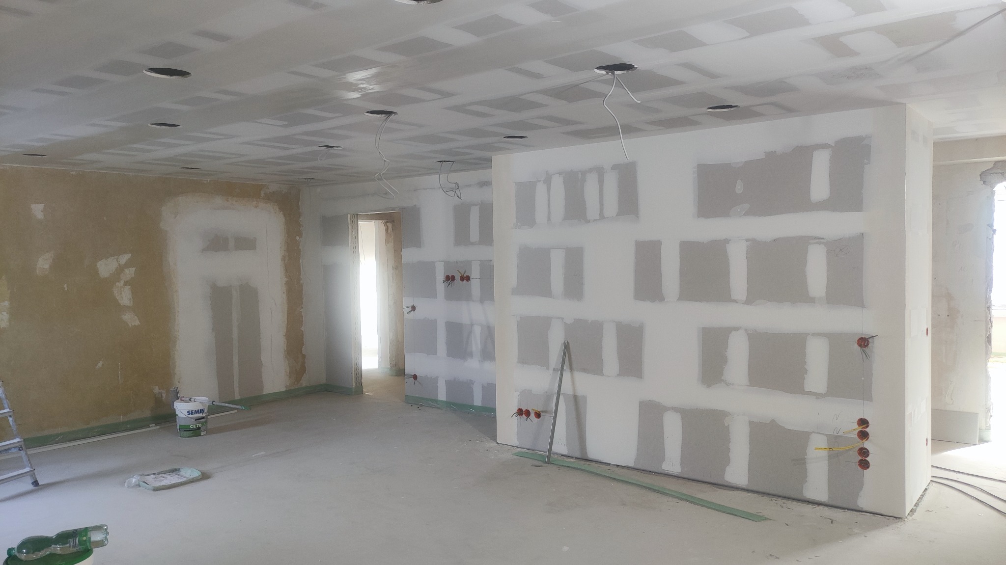 Wnętrze w trakcie montażu płyt kartonowo-gipsowych na ścianach i suficie, widoczne puszki elektryczne i częściowo wykończona ściana z resztkami starej farby, wiadro z gładzią i drabina w tle.
