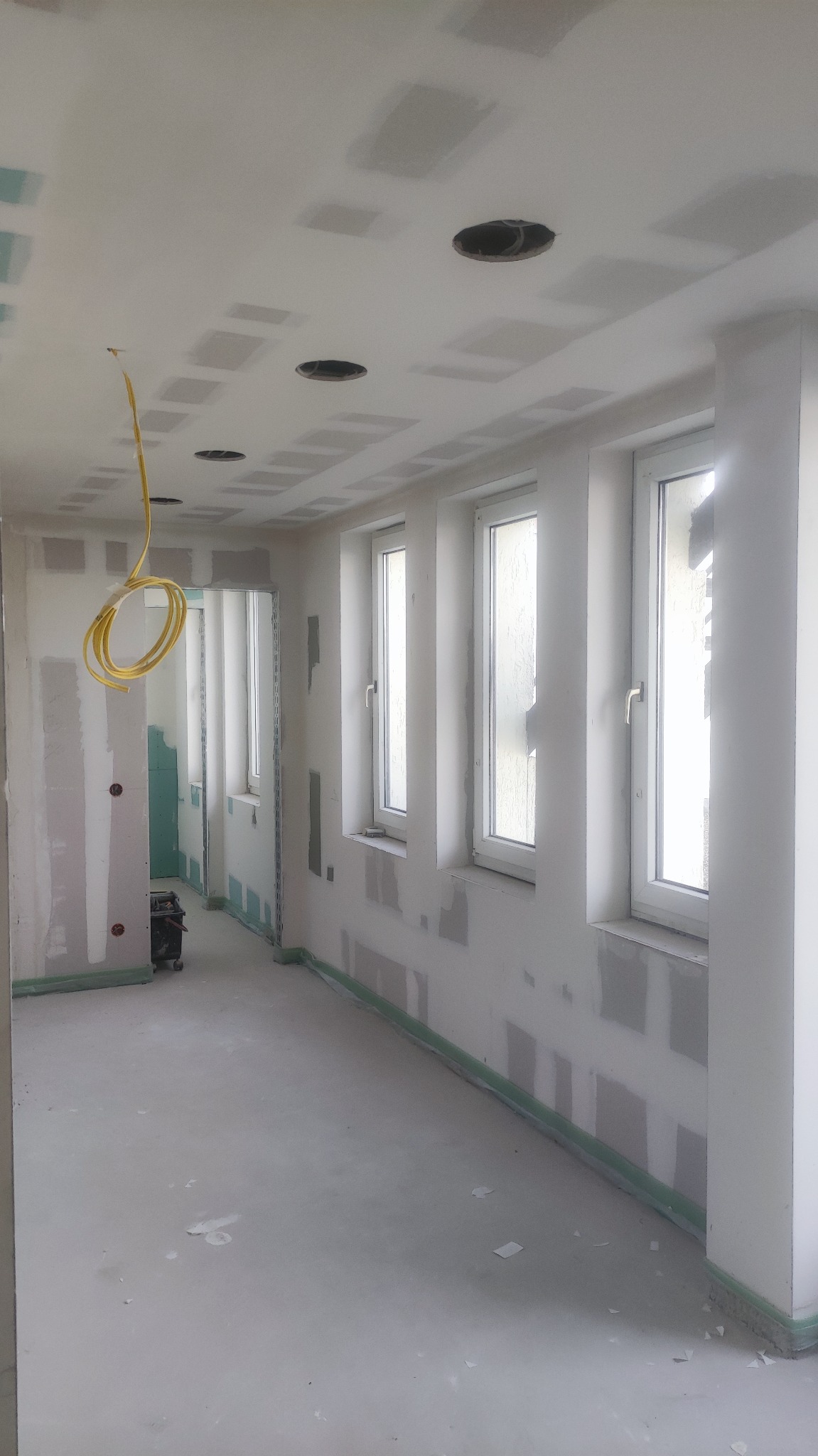 Wnętrze w trakcie remontu: ściany z płyt kartonowo-gipsowych, widoczne łączenia wymagające szpachlowania, sufit z otworami na oświetlenie, zwisający przewód elektryczny, okna z widocznymi...