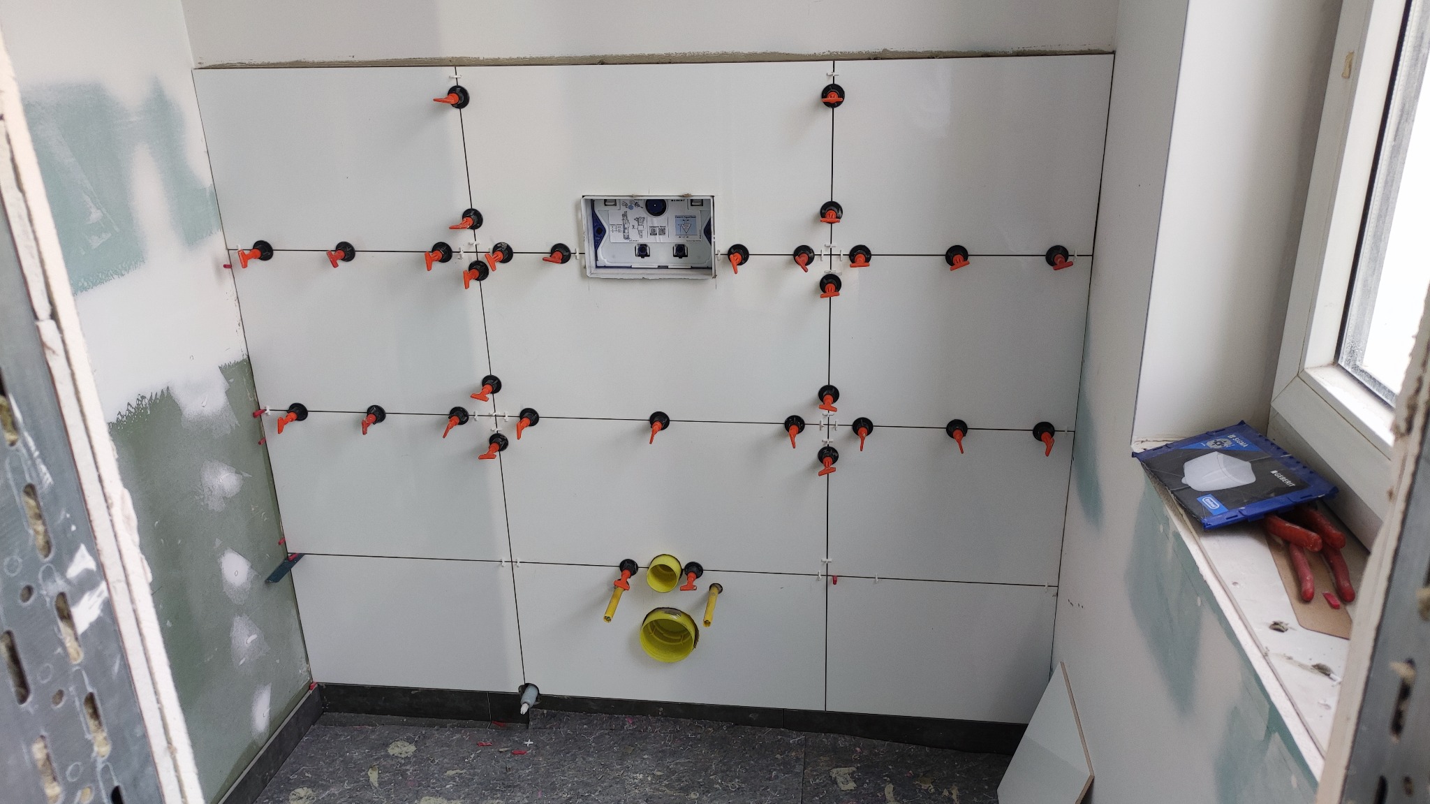 Białe płytki ścienne w trakcie układania w łazience, widoczne krzyżyki dystansowe i poziomica, instalacja podtynkowa w ścianie, fragment ściany z płytami gipsowo-kartonowymi w tle.