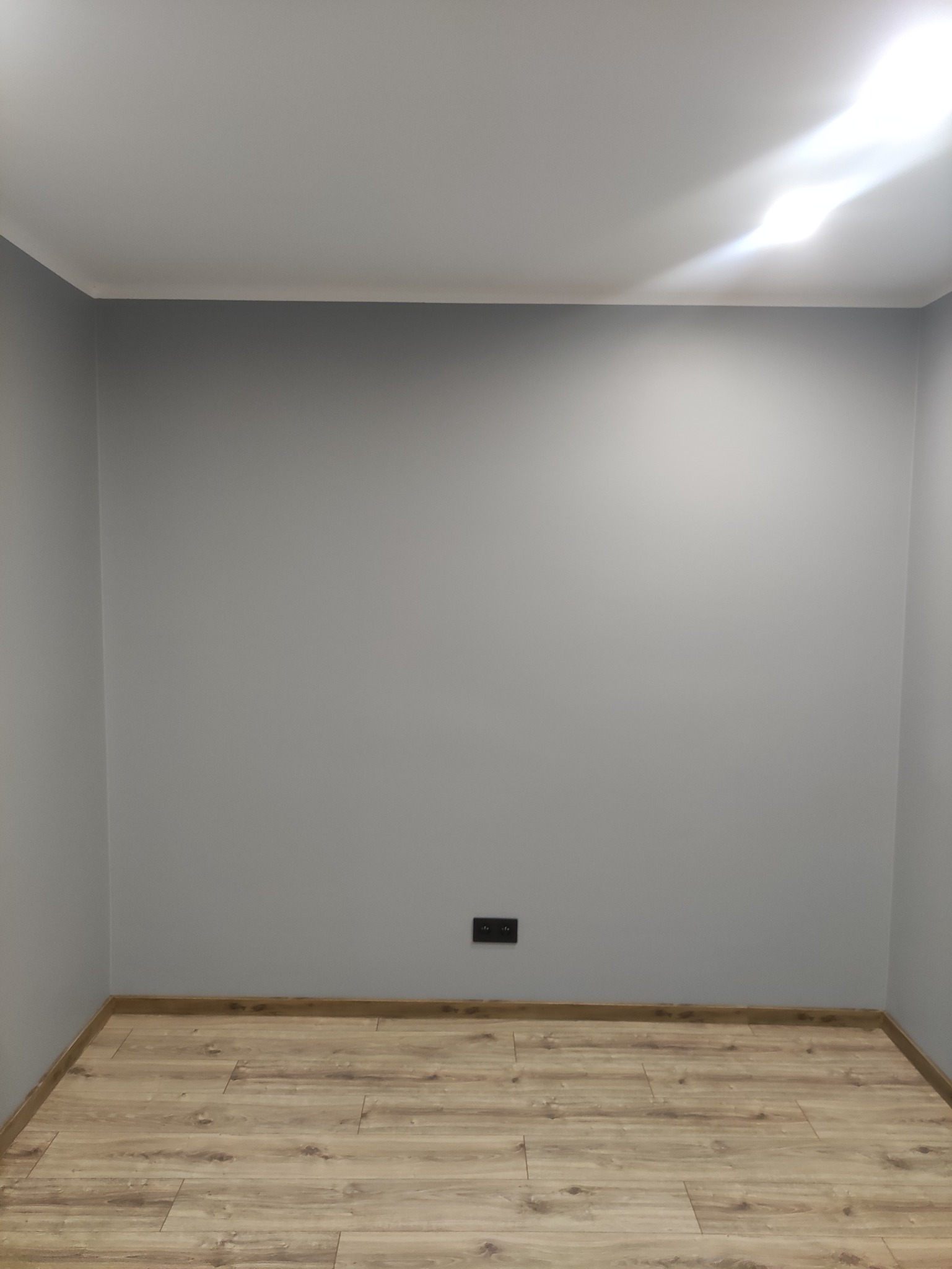 Pomalowany na szaro pokój z ułożonymi panelami podłogowymi i gładkim sufitem, widoczne gniazdko elektryczne.