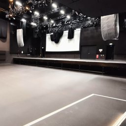 PAWEŁ GUNIA PAGAN - Szara, gładka posadzka w dużej sali widowiskowej z oświetleniem technicznym. Widoczne białe linie na podłodze i czarne ściany. Scena z ekranem w tle.