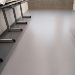 PAWEŁ GUNIA PAGAN - Gładka, szara posadzka w jasnym pomieszczeniu z oknami i betonową ścianą. Widoczne fragmenty metalowych nóg stołów. Minimalistyczny, nowoczesny design.