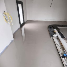 PAWEŁ GUNIA PAGAN - Gładka, szara posadzka w surowym wnętrzu z widocznymi przewodami i narzędziami, świeżo po wylaniu, z czarną ramą drzwi w tle. Minimalistyczny, nowoczesny styl.