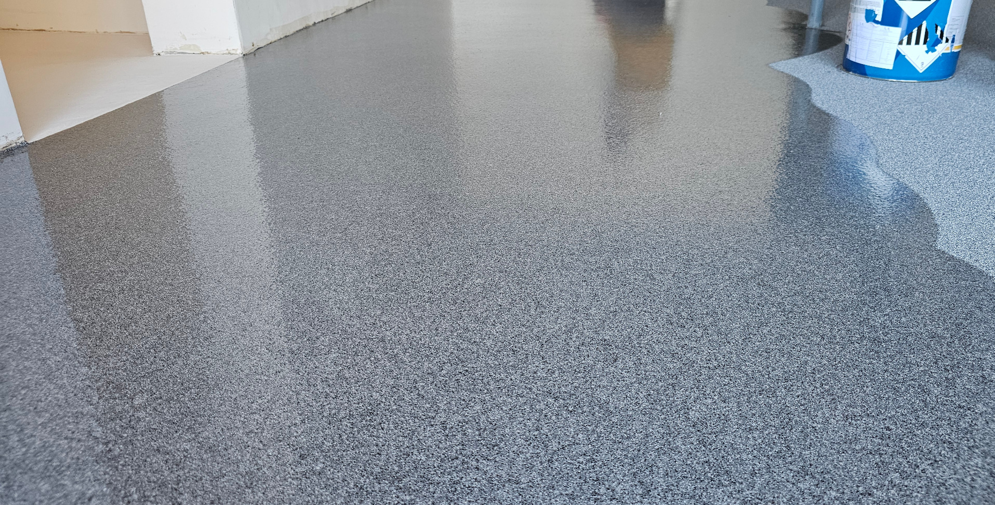 Błyszcząca, szara posadzka żywiczna z widoczną teksturą i refleksami światła. W tle ściana i wiadro z farbą, co wskazuje na świeżo wykonaną realizację.