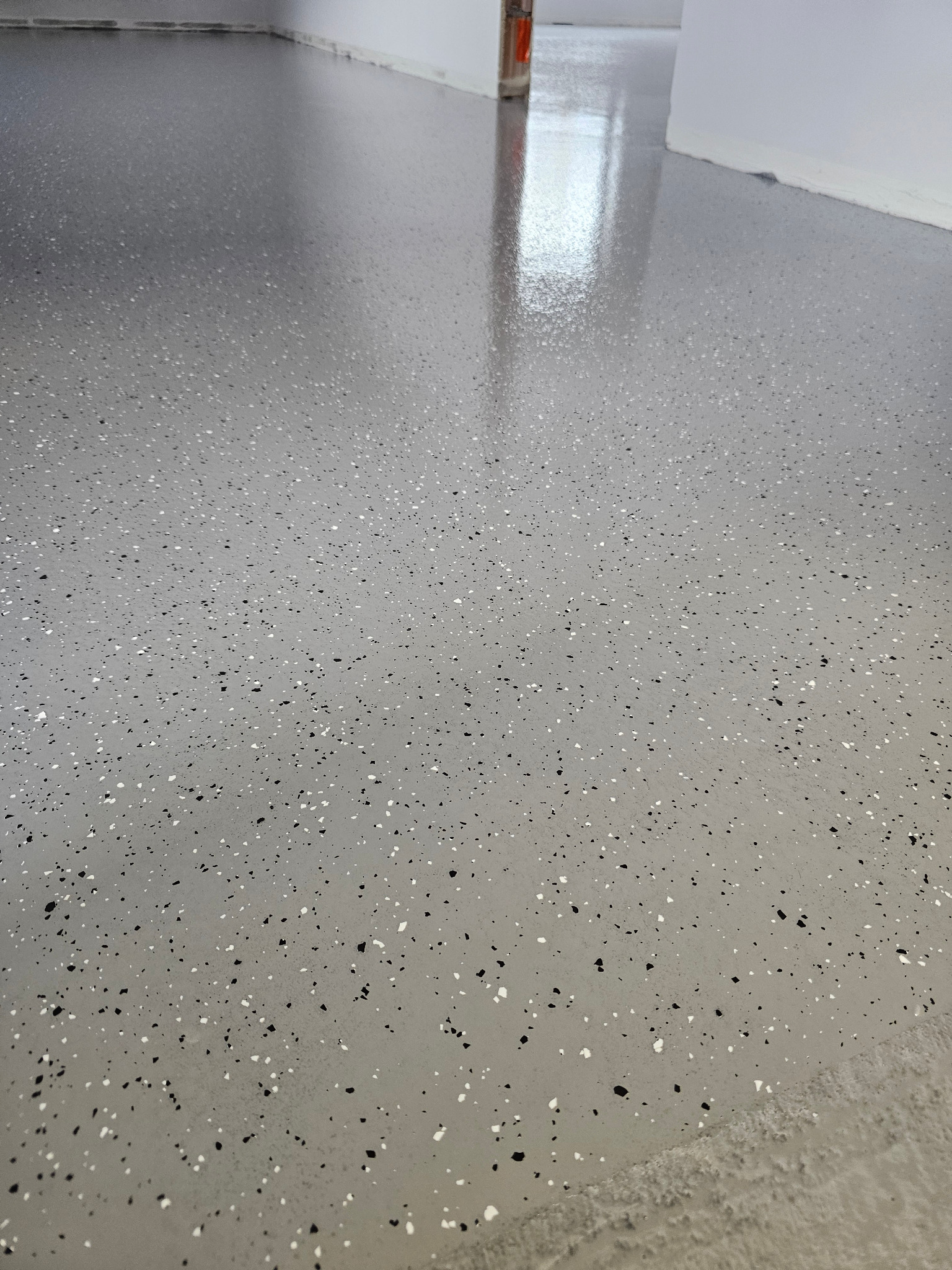 Gładka, szara powierzchnia podłogi z żywicy z czarnymi i białymi drobinkami. Widoczne odbicie światła, podkreślające błyszczące wykończenie. Minimalistyczny design.