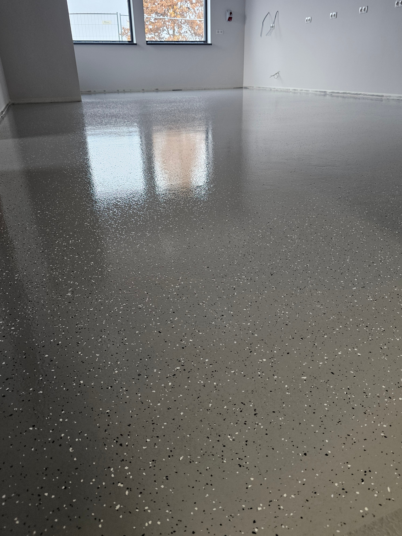 Błyszcząca, szara posadzka z żywicy epoksydowej z czarnymi i białymi drobinkami w nowym, jasnym pomieszczeniu z oknami i gniazdkami elektrycznymi na ścianie.