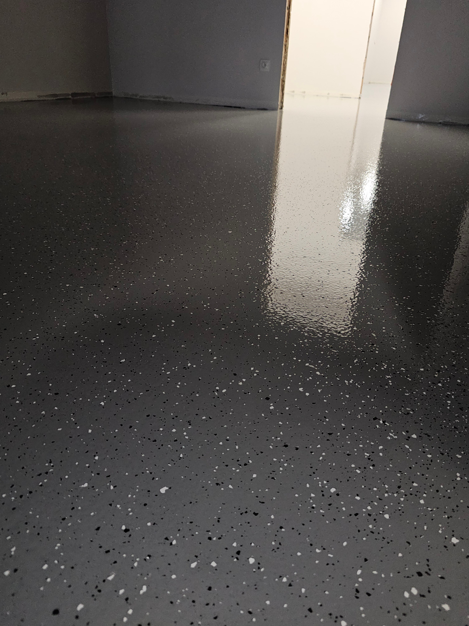 Gładka, szara posadzka z białymi i czarnymi drobinami, odbijająca światło. Widoczne fragmenty ścian i futryny drzwi.
