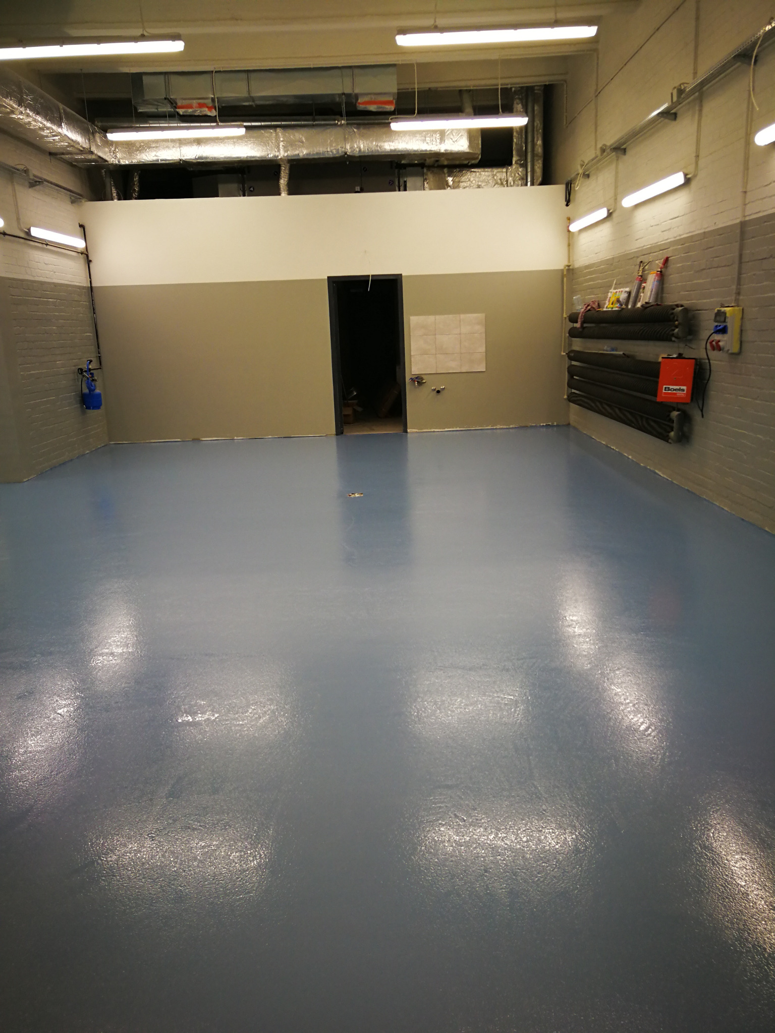 Błękitna, gładka posadzka w przemysłowym wnętrzu z szaro-białymi ścianami. Widoczne rury pod sufitem i grzejniki na ścianie, oświetlenie jarzeniowe.