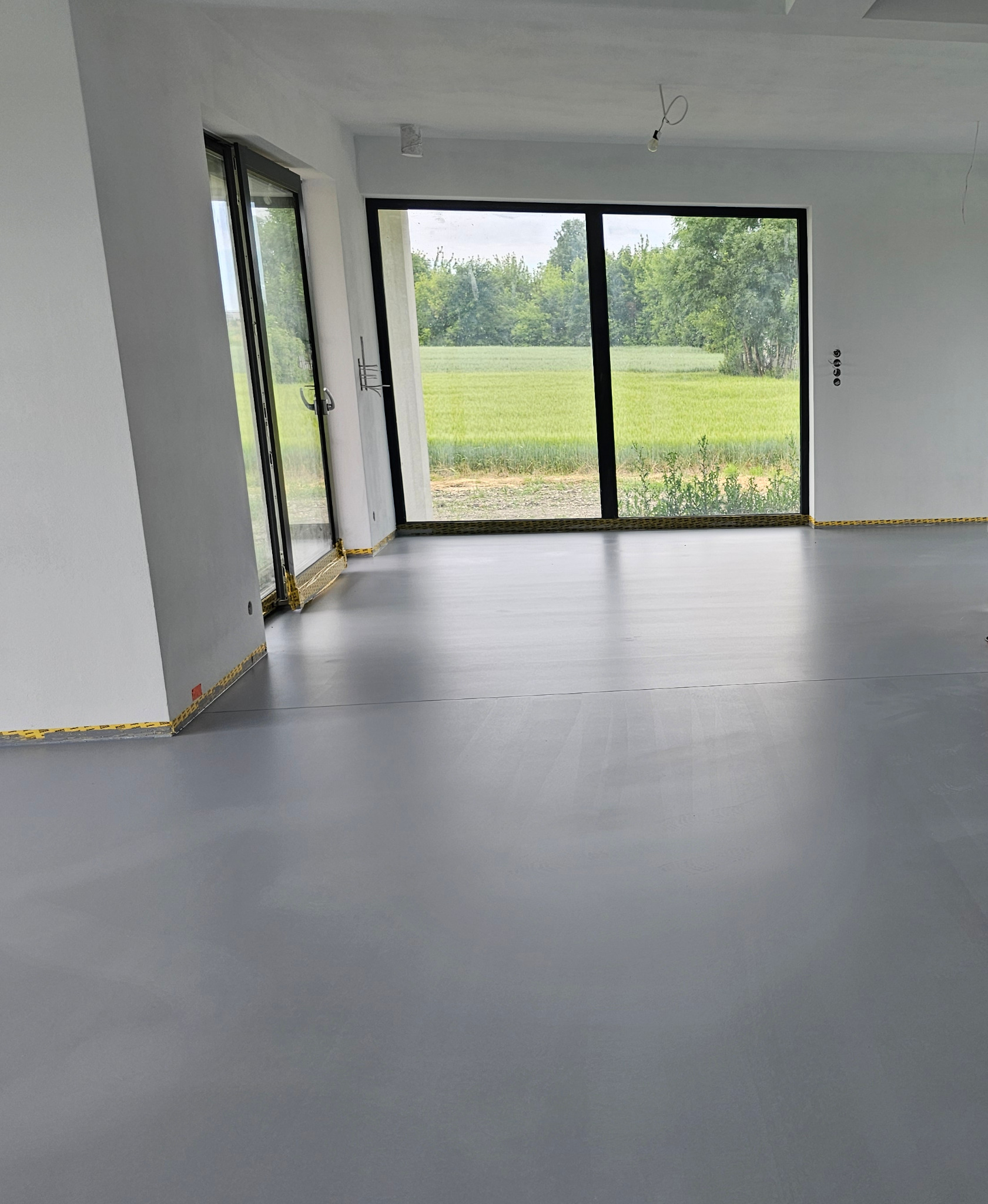 Szara, błyszcząca posadzka w nowoczesnym wnętrzu z dużymi oknami na zielony krajobraz. Taśma zabezpieczająca przy ścianach. Minimalistyczny design.