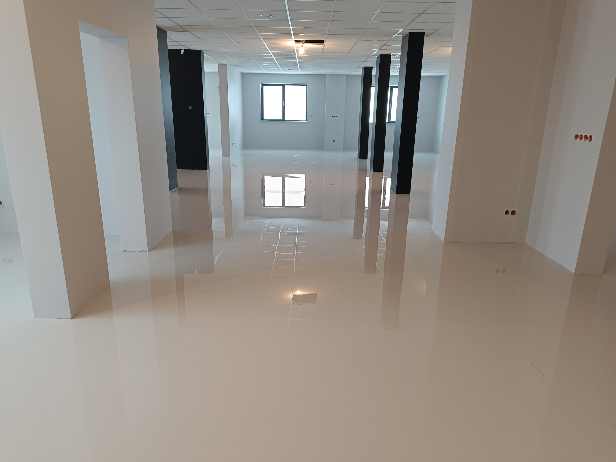 Błyszcząca, jasna posadzka w przestronnym wnętrzu biurowym z białymi i czarnymi kolumnami. Widoczne okno i odbicia światła na gładkiej powierzchni.