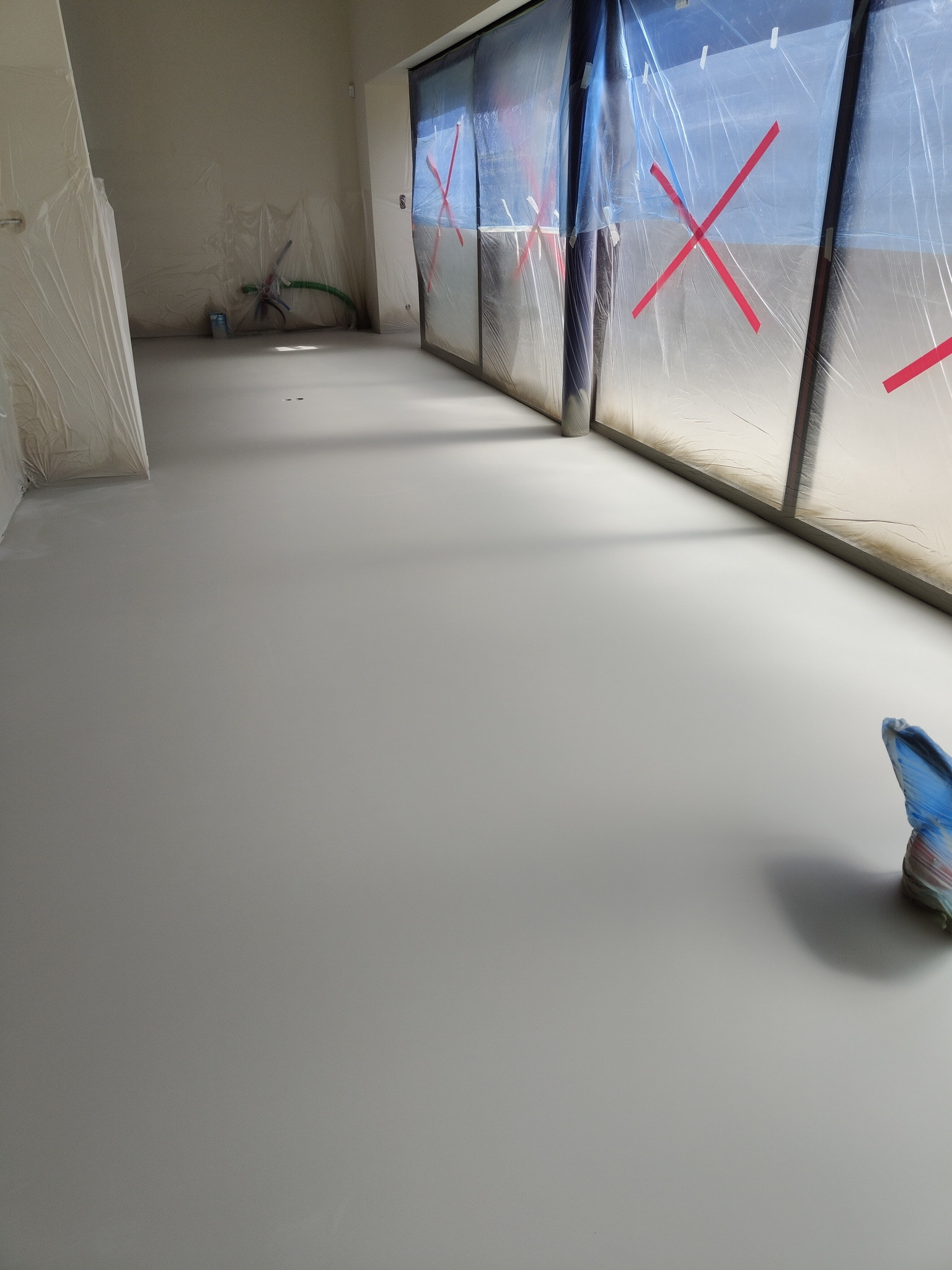 Gładka, szara powierzchnia w pomieszczeniu po wylaniu posadzki. Okna zabezpieczone folią malarską. Widoczny fragment narzędzia do rozprowadzania masy.