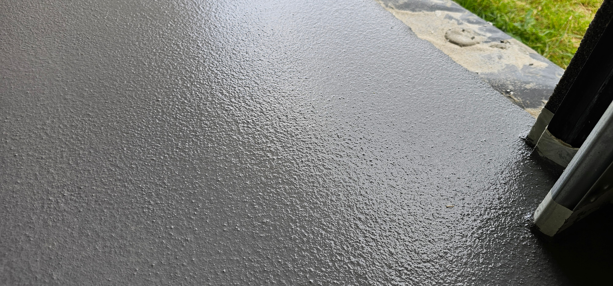 Świeżo wylana, szara posadzka z żywicy epoksydowej, z widocznym narzędziem do rozprowadzania. Delikatna faktura powierzchni, fragment zieleni w tle.