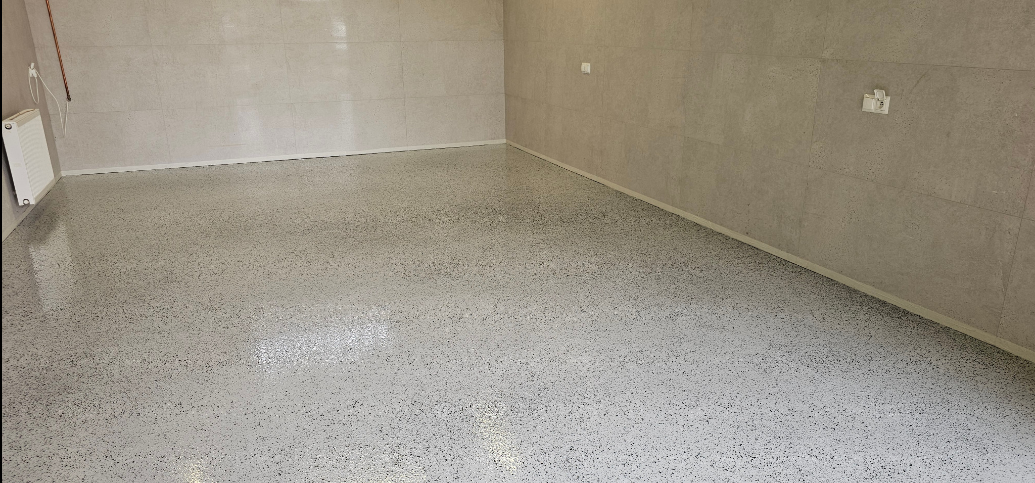 Jasna, gładka posadzka z drobnym wzorem w pomieszczeniu wyłożonym płytkami. Widoczny grzejnik i gniazdko elektryczne. Nowoczesny minimalizm.