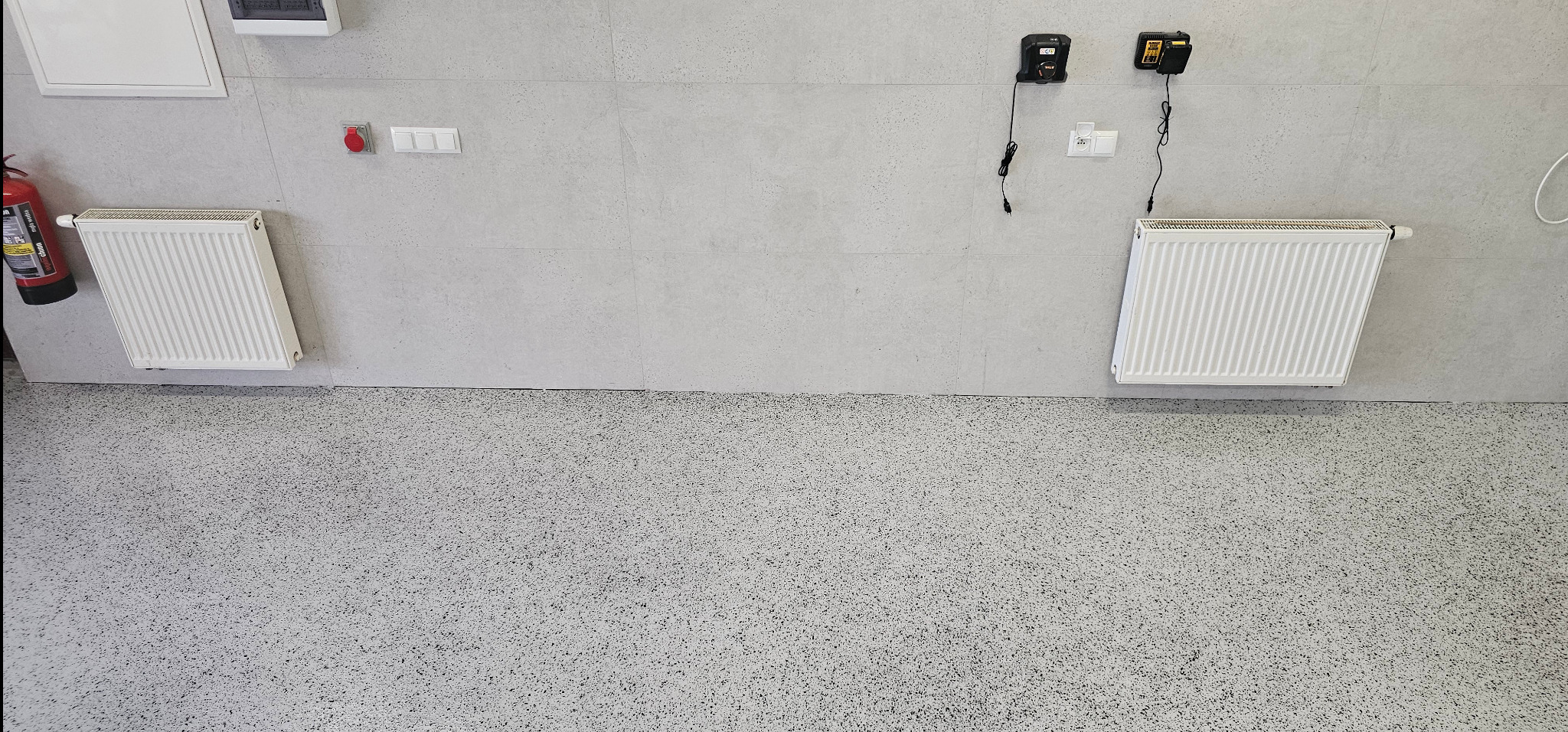 Jasnoszara posadzka żywiczna z widocznymi grzejnikami i elementami instalacji elektrycznej na ścianie. Widok z góry na gładką powierzchnię.