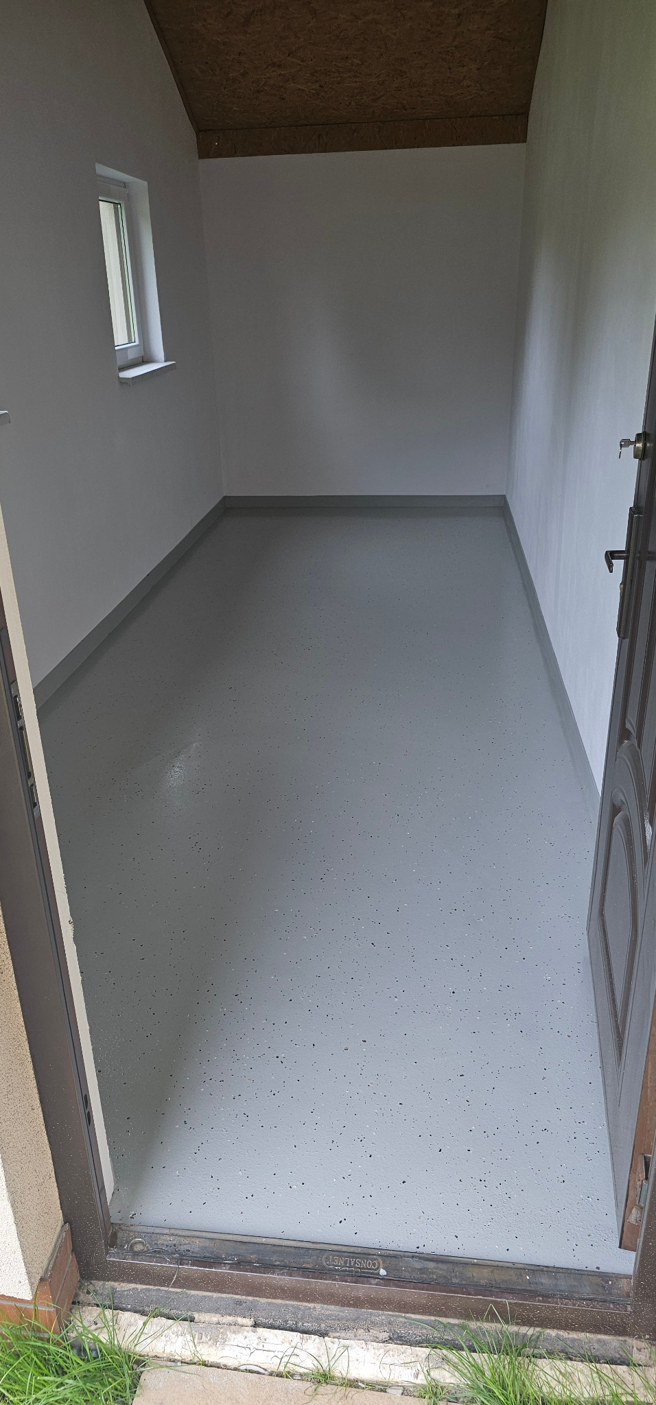 Szara posadzka żywiczna z ciemnymi drobinkami w pomieszczeniu z białymi ścianami, widok z progu przez otwarte drzwi. Małe okno po lewej stronie.