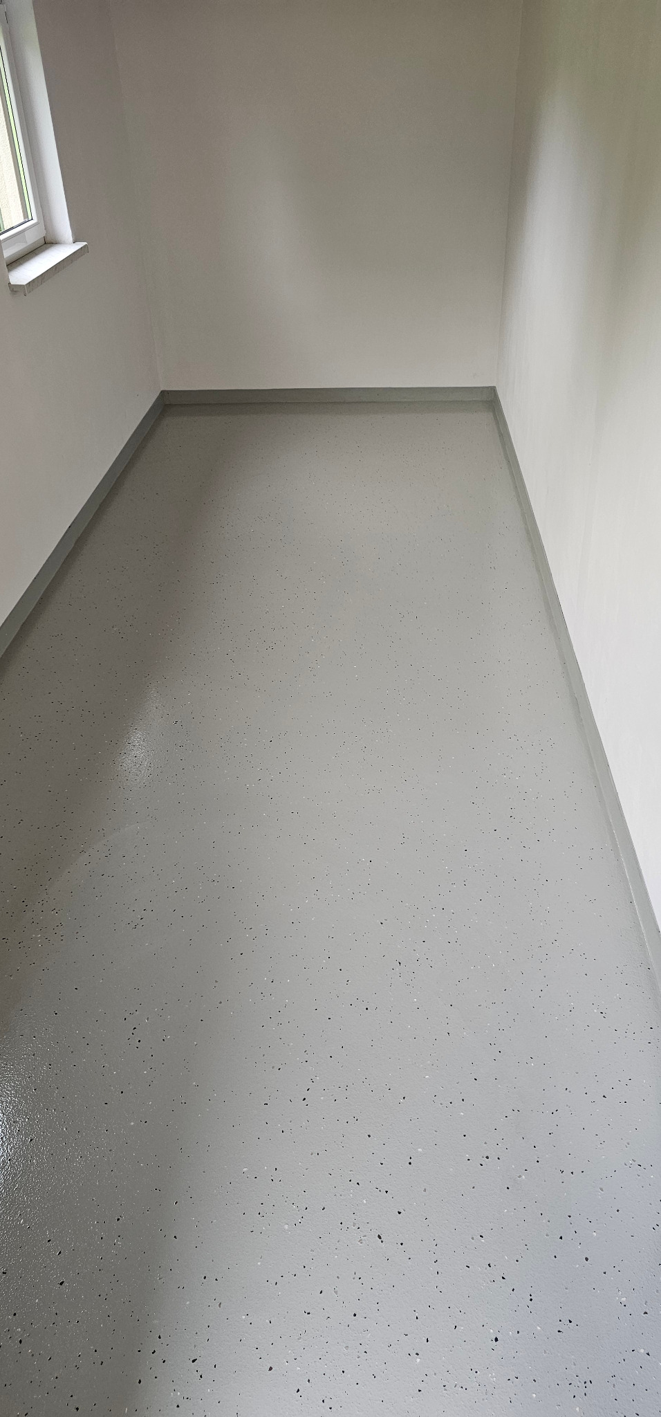 Szara, gładka posadzka z drobnymi, ciemnymi wtrąceniami w narożniku pomieszczenia, widok z góry. Minimalistyczne wykończenie wnętrza, białe ściany, okno w lewym górnym rogu.