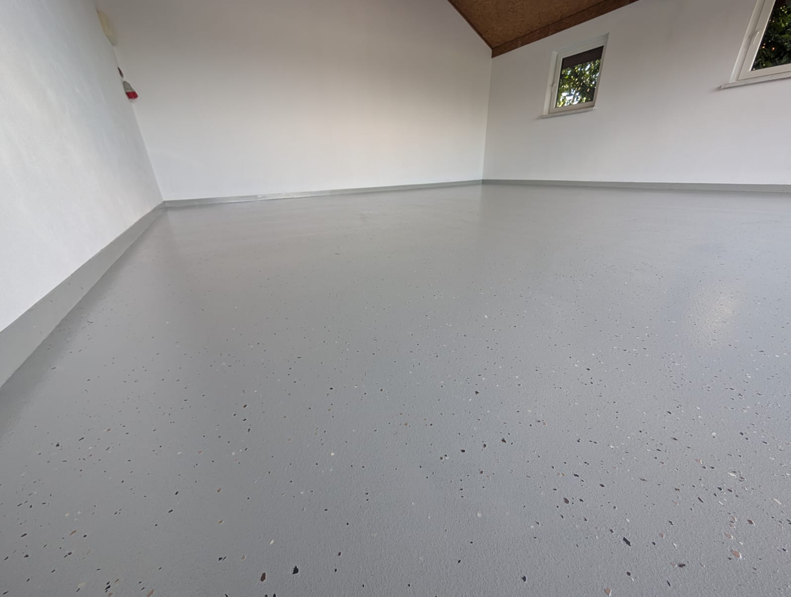 Jasnoszara, gładka posadzka z delikatnym, wielobarwnym wzorem w garażu. Widoczne okna i białe ściany, tworzące czyste i nowoczesne wnętrze.