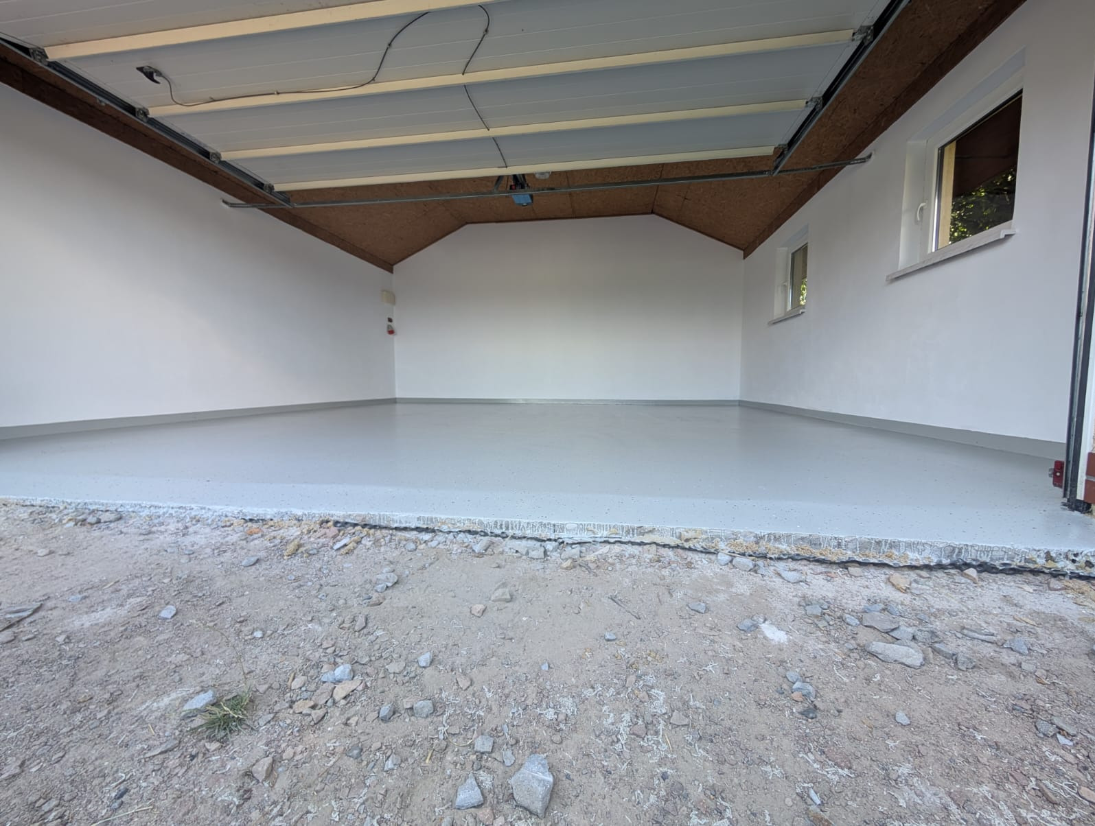 Jasnoszara, gładka posadzka w garażu z białymi ścianami i oknami. Widoczny fragment podsufitki i surowy grunt przed wjazdem, podkreślający świeżość wykonania.