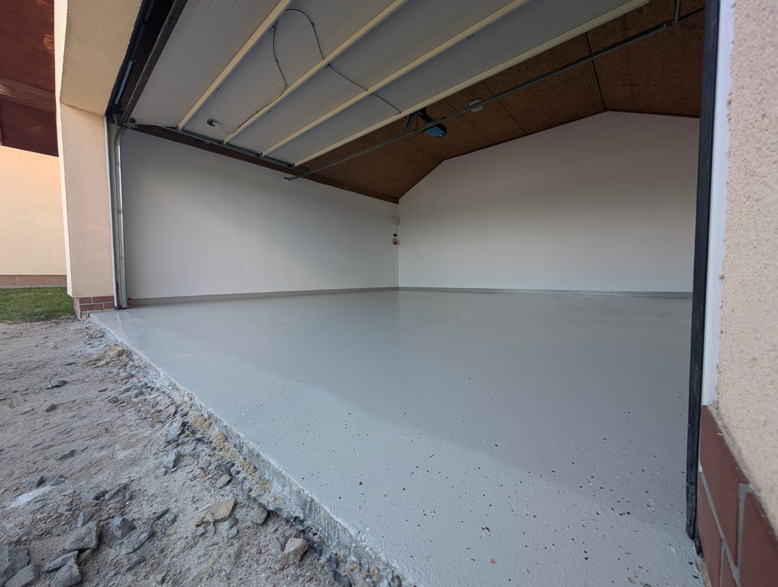 Jasnoszara, gładka posadzka w garażu z otwartą bramą. Widoczne drobne, ciemne plamki w strukturze. Ściany białe, sufit z widoczną konstrukcją.