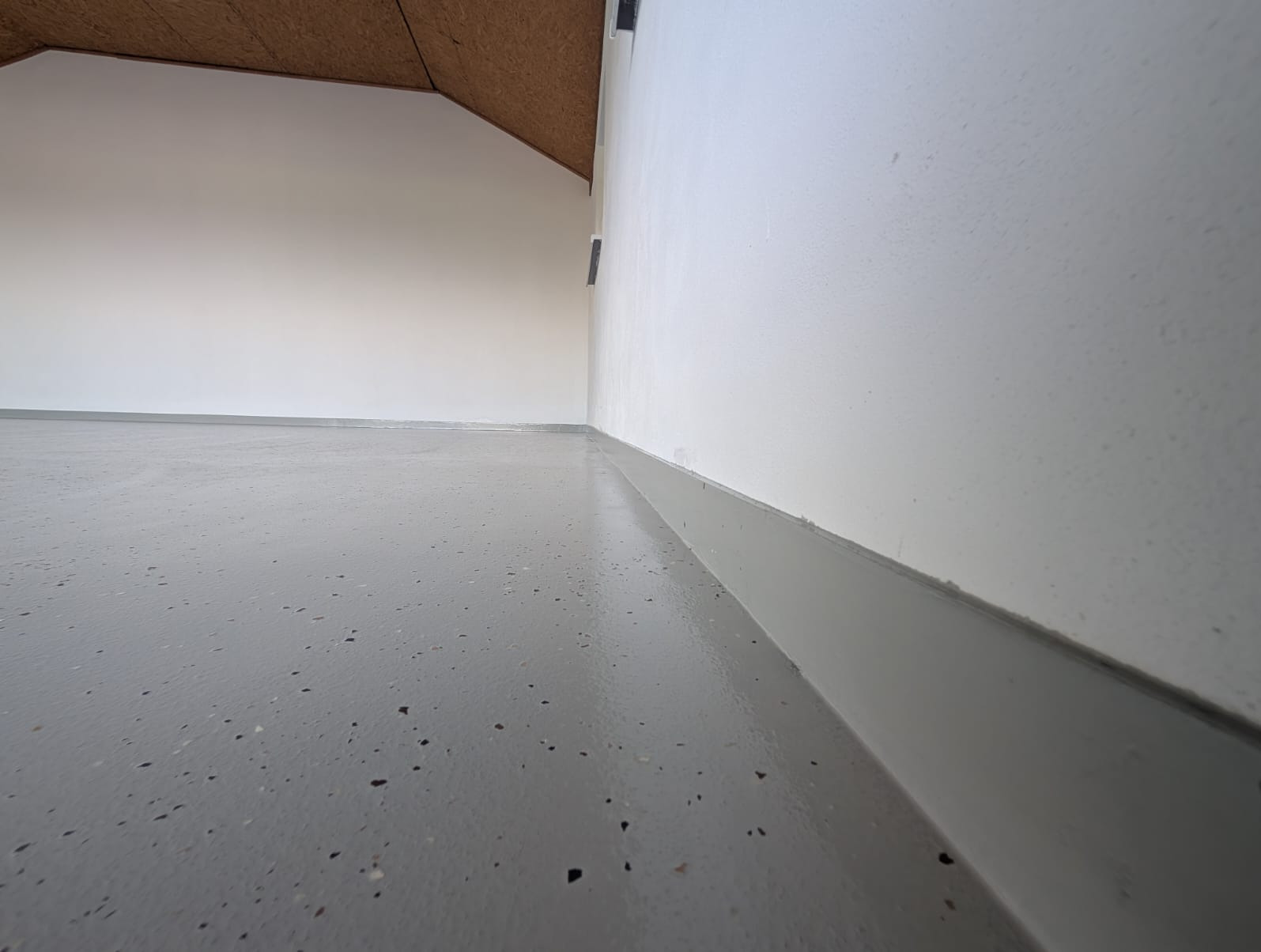 Szara, błyszcząca posadzka żywiczna z ciemnymi inkluzjami, świeżo wylana w pomieszczeniu ze skośnym stropem w Krakowie. Widok z niskiej perspektywy, oświetlenie naturalne.