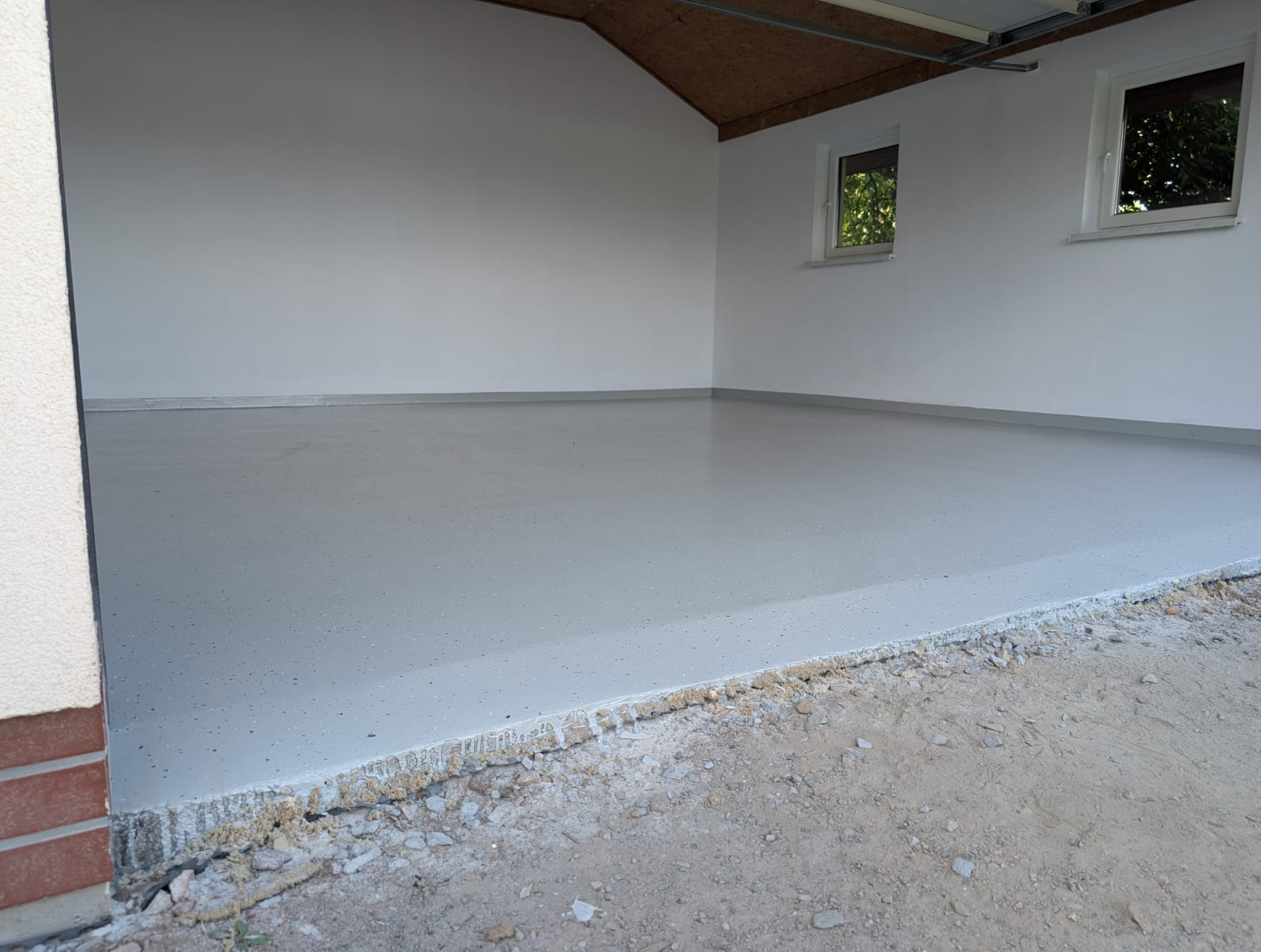 Jasnoszara, gładka powierzchnia w garażu po nałożeniu posadzki. Widoczne okna i sufit, stan surowy przed wykończeniem. Krawędź posadzki przy wejściu do garażu.