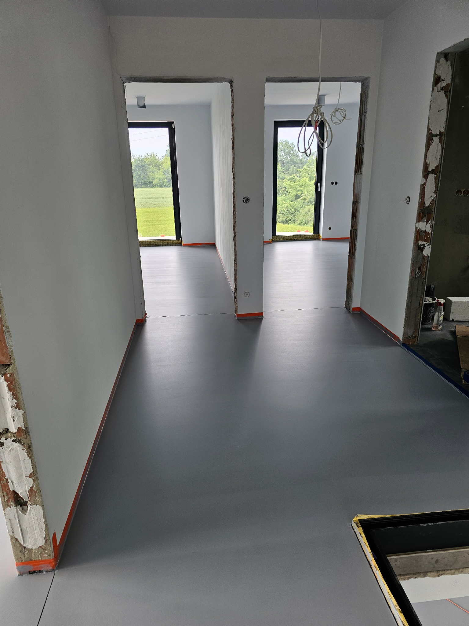 Szara posadzka żywiczna w jasnym wnętrzu z widokiem na zieleń przez okna. Minimalistyczny design, surowe ściany, wykończone wylewki, brak listew przypodłogowych.