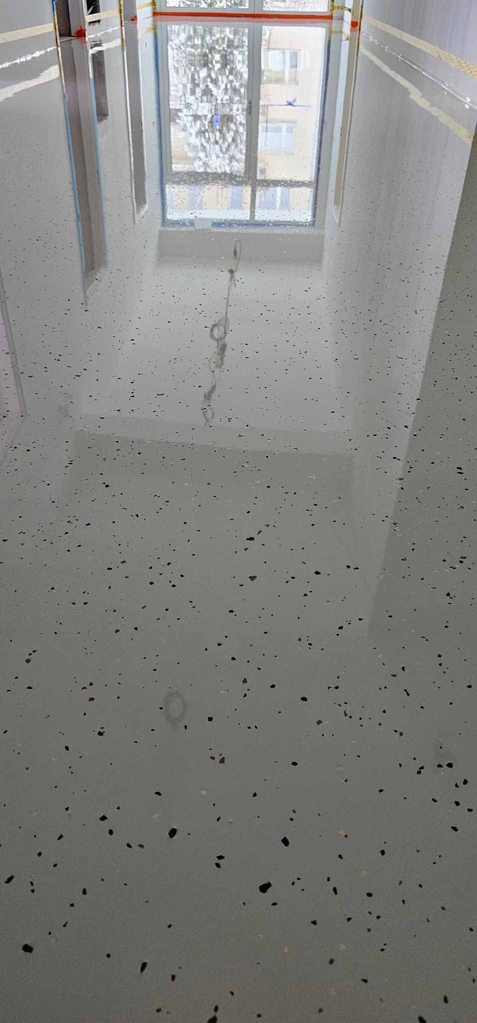 Jasnoszara, błyszcząca posadzka z ciemnymi, nieregularnymi plamkami, odbijająca okno. Widok z góry, ukazujący gładką powierzchnię i wzór.