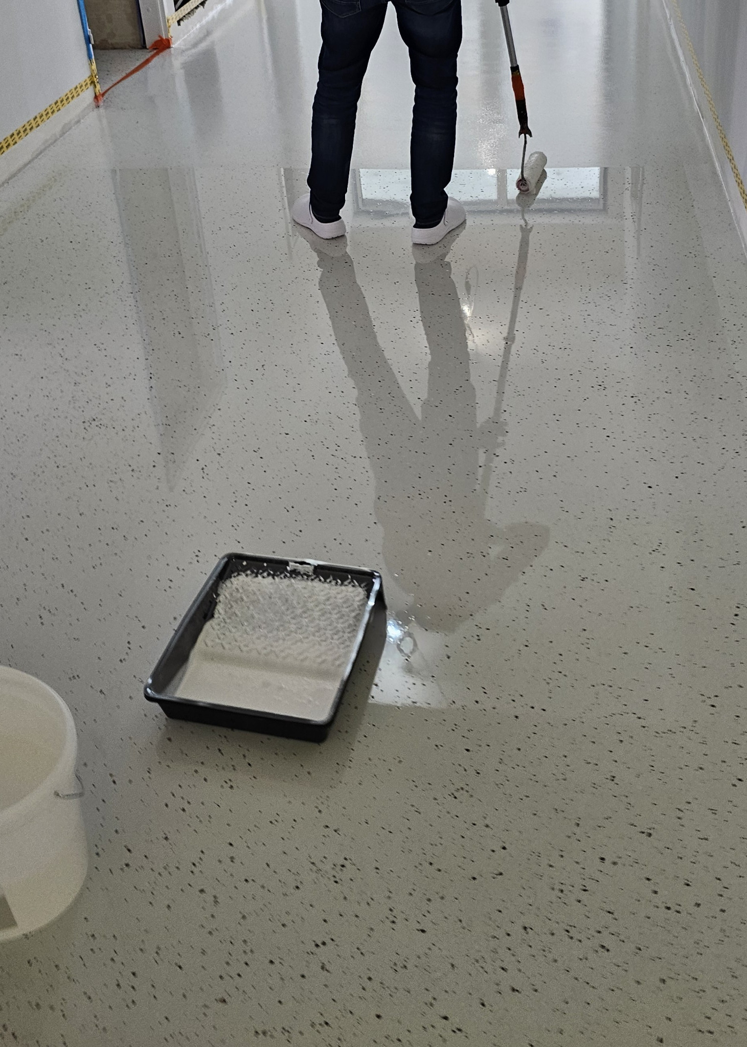 Aplikacja jasnej, błyszczącej posadzki z drobnymi, ciemnymi inkluzjami za pomocą wałka. Widoczny kuweta z farbą i częściowo wiadro.