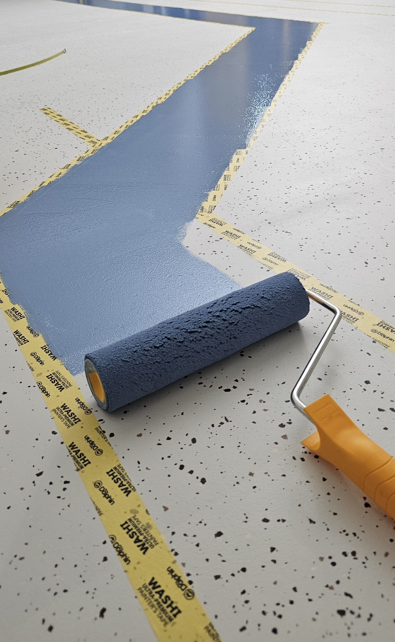 Malowanie podłogi wałkiem na szaro, wyznaczona taśmą malarską. Widoczna struktura podłoża z drobnymi plamkami. Detal wałka i taśmy malarskiej.