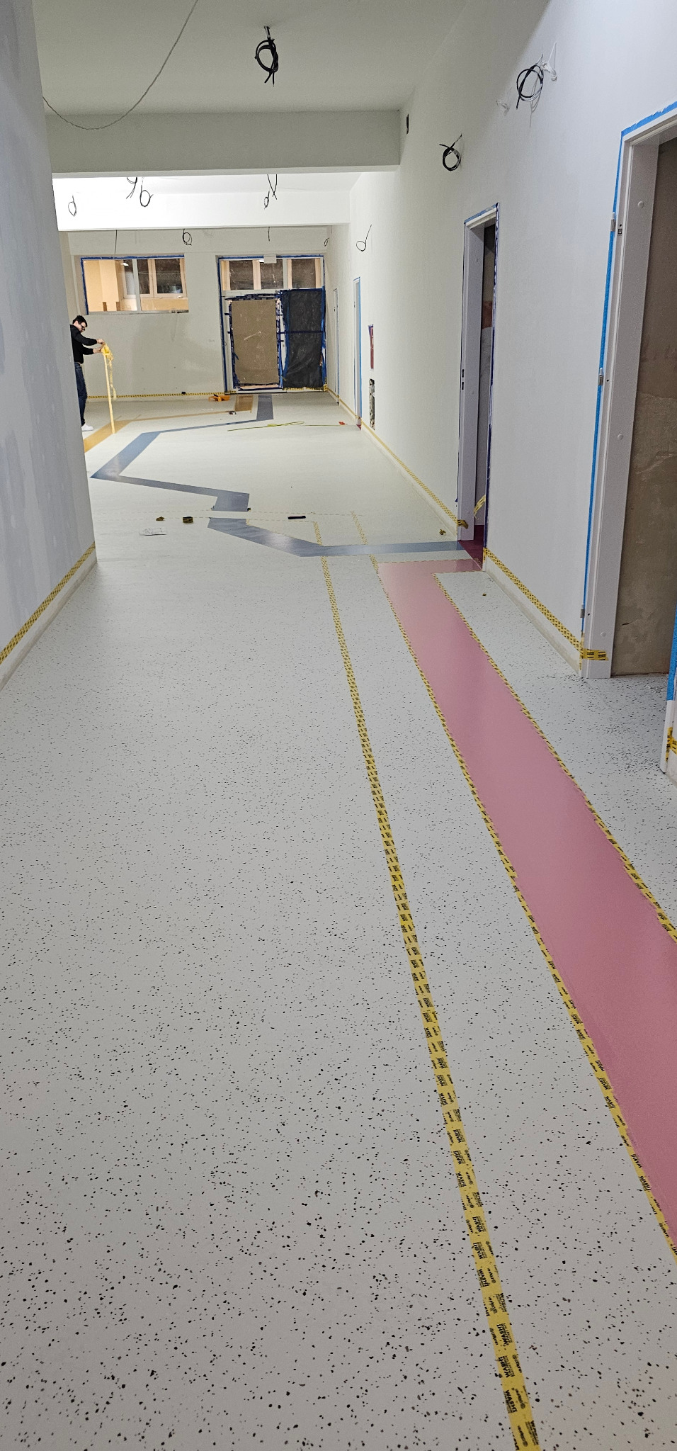 Wnętrze w trakcie prac wykończeniowych: jasna posadzka z ciemnymi plamkami, zabezpieczona taśmą malarską, widoczne instalacje pod sufitem i fragment osoby w oddali.