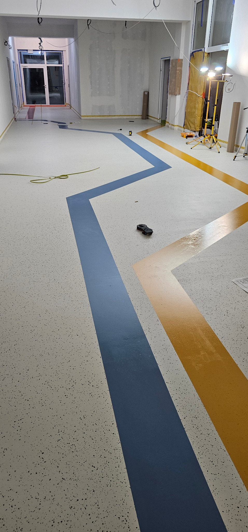 Posadzka żywiczna z dekoracyjnymi pasami w kolorze niebieskim i żółtym, ułożonymi w zygzak. Widoczny laserowy niwelator i lampy oświetleniowe.