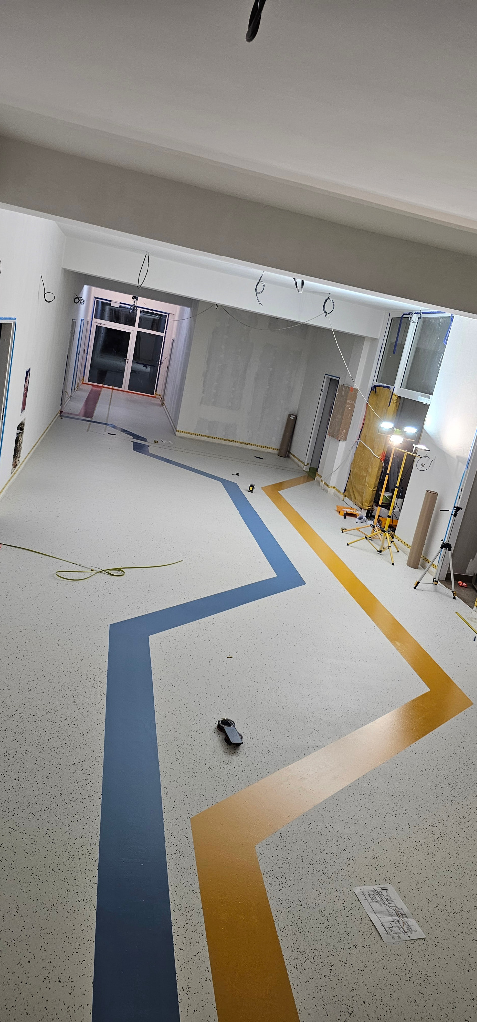 Wnętrze w trakcie wykańczania z wzorem zygzak na posadzce w kolorach niebieskim i żółtym. Widoczne narzędzia i drabina. Jasne oświetlenie.