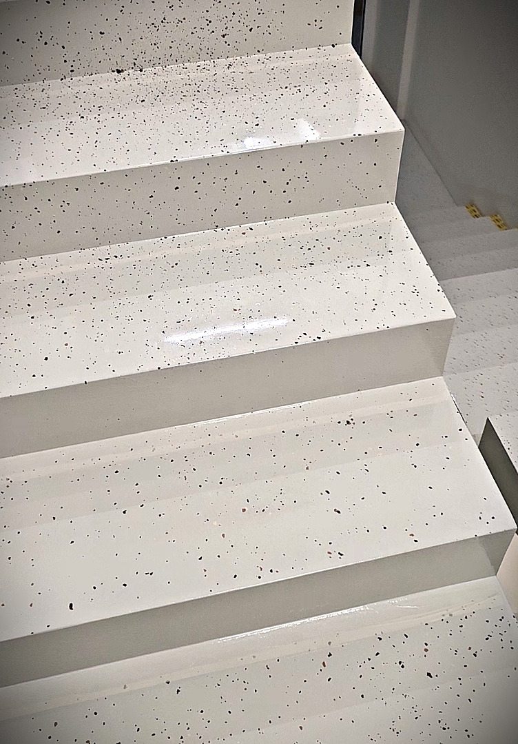 Schody wykończone jasną, błyszczącą powierzchnią z ciemnymi, drobnymi plamkami. Widok z góry, ukazujący kilka stopni. Minimalistyczny design.