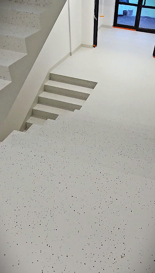 Jasnoszara posadzka z drobnymi, ciemnymi plamkami, schody z tego samego materiału. Widok z góry, w tle okno i drzwi.