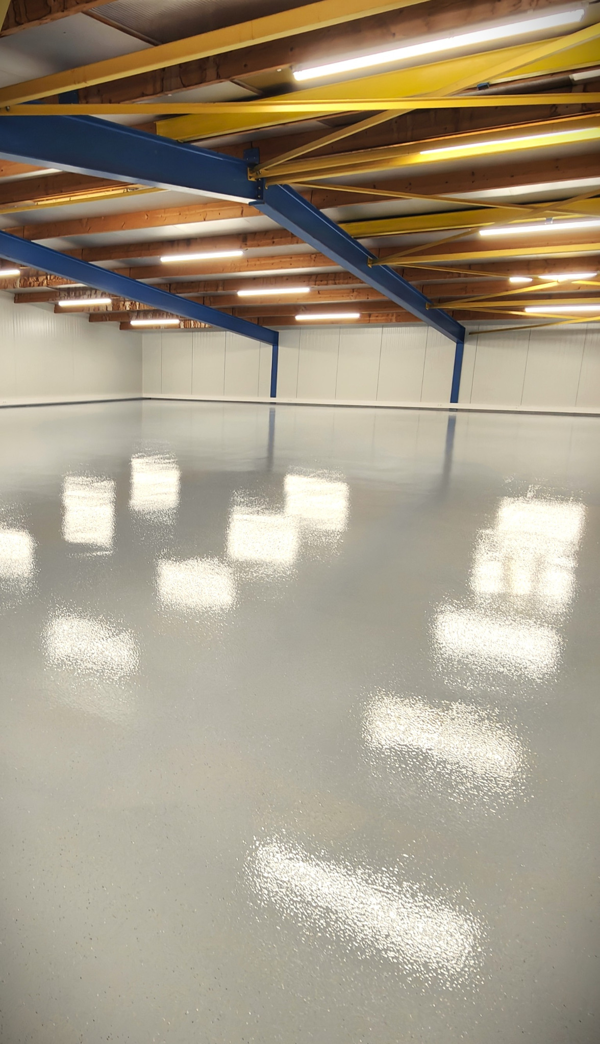 Jasnoszara, błyszcząca posadzka w hali przemysłowej z widoczną konstrukcją stalową pod sufitem i panelami ściennymi. Odbicia światła na gładkiej powierzchni.