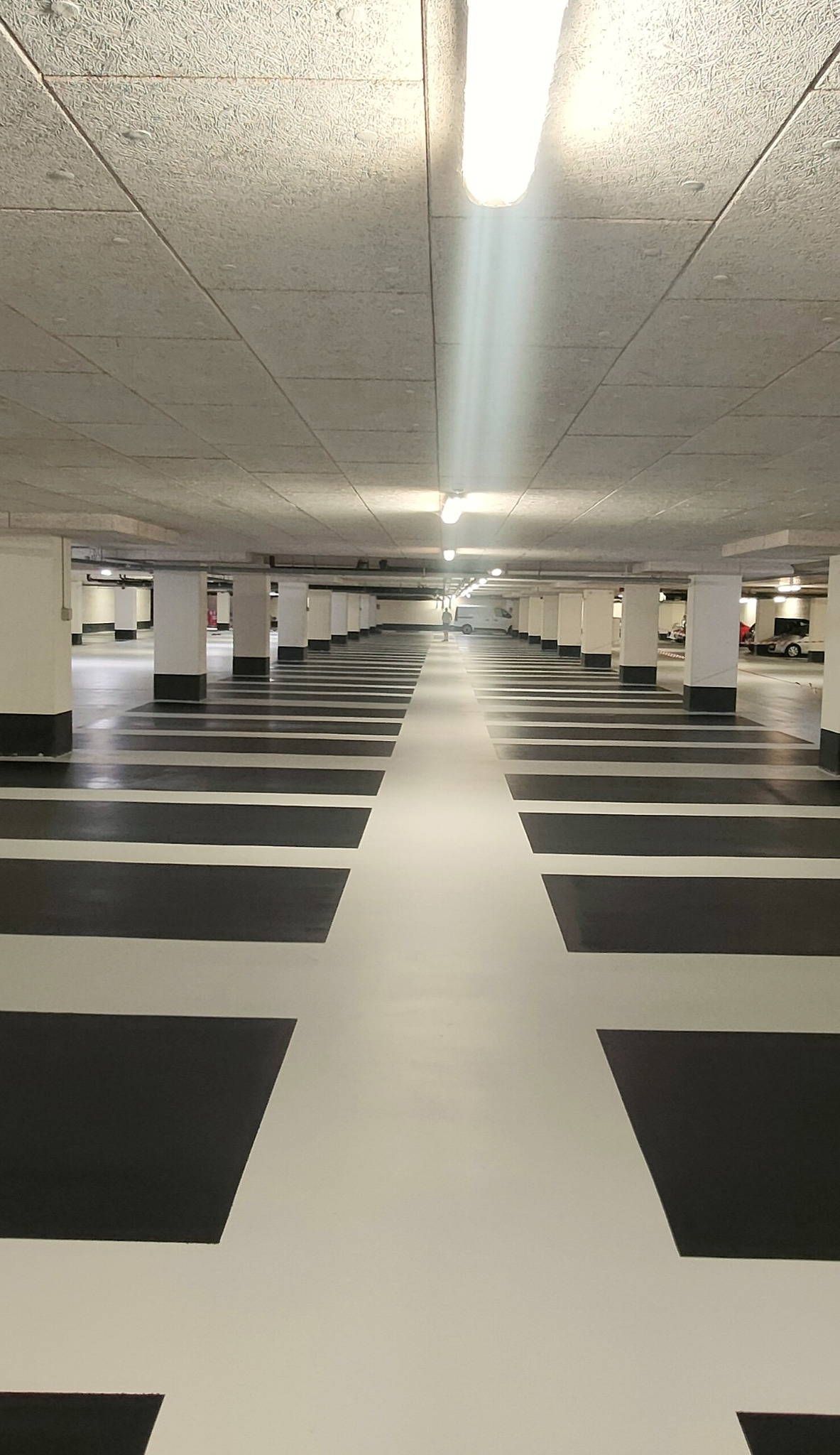 Biało-czarna posadzka w garażu podziemnym z rzędami kolumn. Perspektywa zbieżna, oświetlenie jarzeniowe. Widoczne samochody w oddali. Gładka powierzchnia.