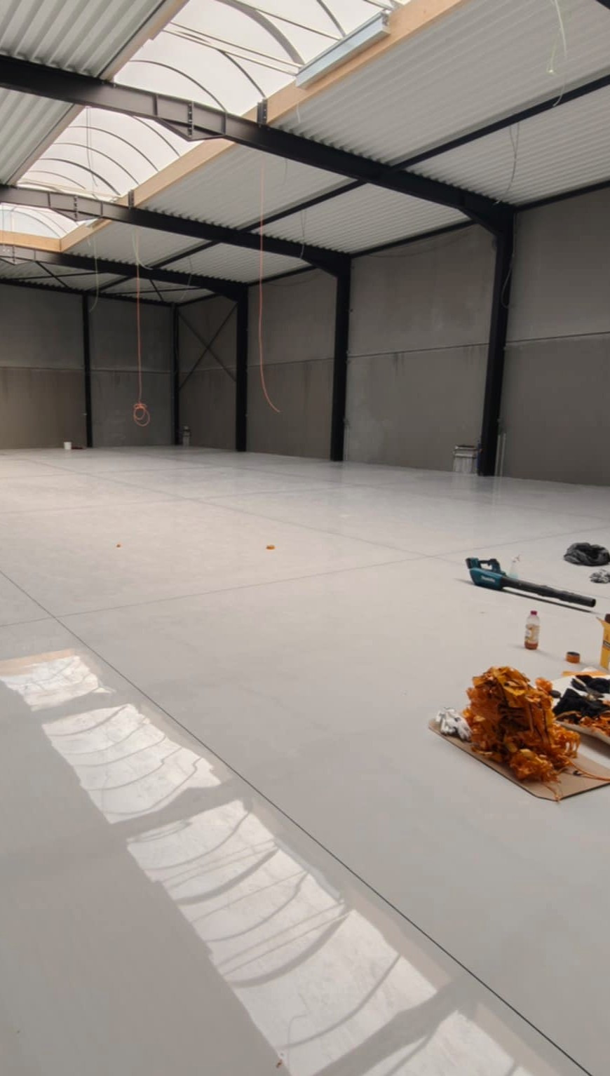 Biała, błyszcząca posadzka żywiczna w hali przemysłowej z widocznymi narzędziami i materiałami budowlanymi. Światło dzienne wpada przez okna dachowe.