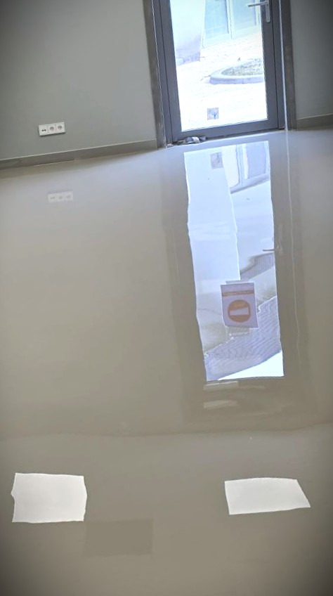 Błyszcząca, jasnoszara podłoga w pokoju z widocznym oknem i odbiciami światła. Gładka powierzchnia, minimalistyczny design, świeżo wykończone wnętrze.