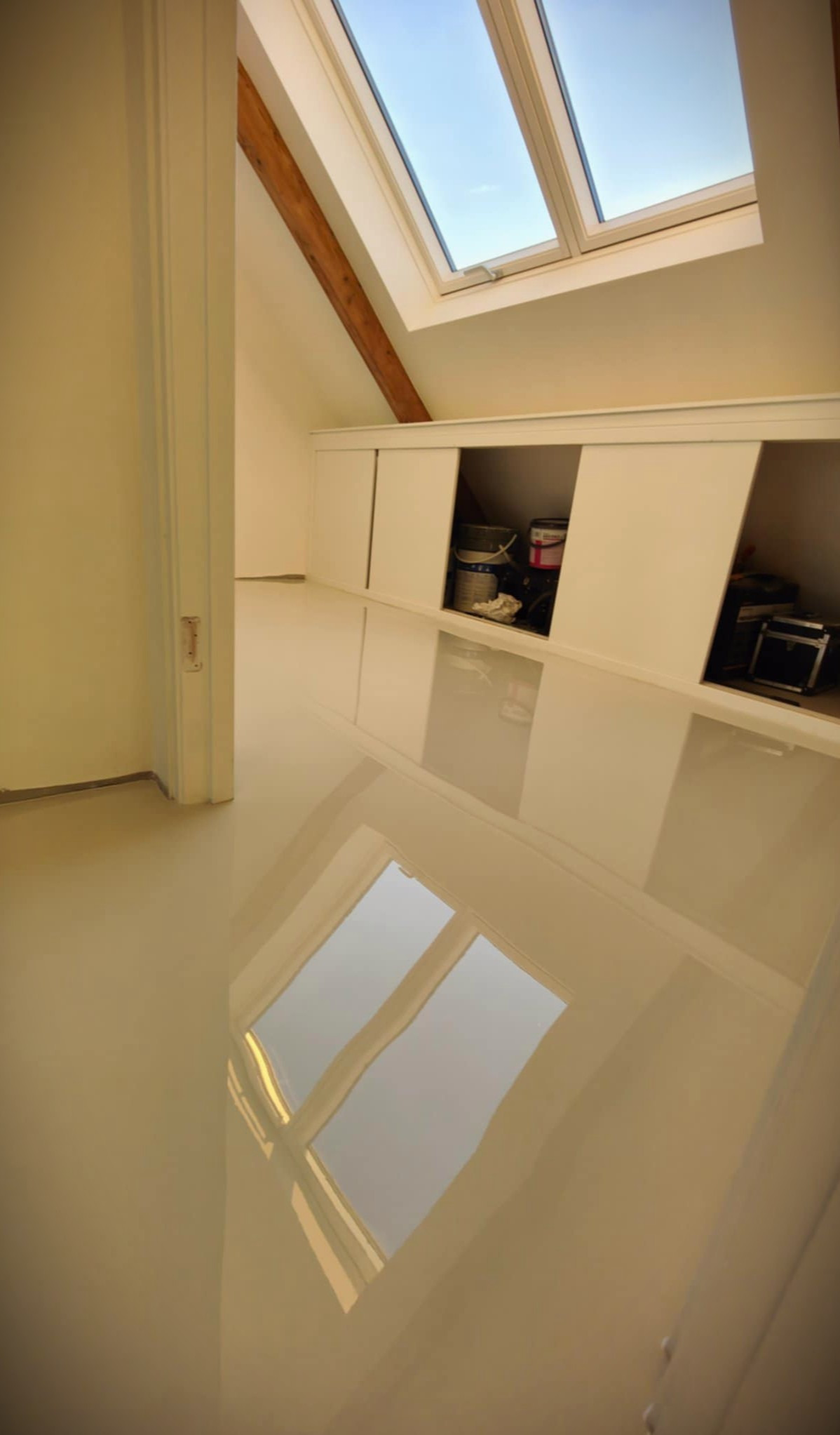Beżowa, lśniąca podłoga w pokoju na poddaszu z oknem dachowym. Gładka powierzchnia odbija światło, tworząc efekt lustra. Widoczne wnęki w ścianie.