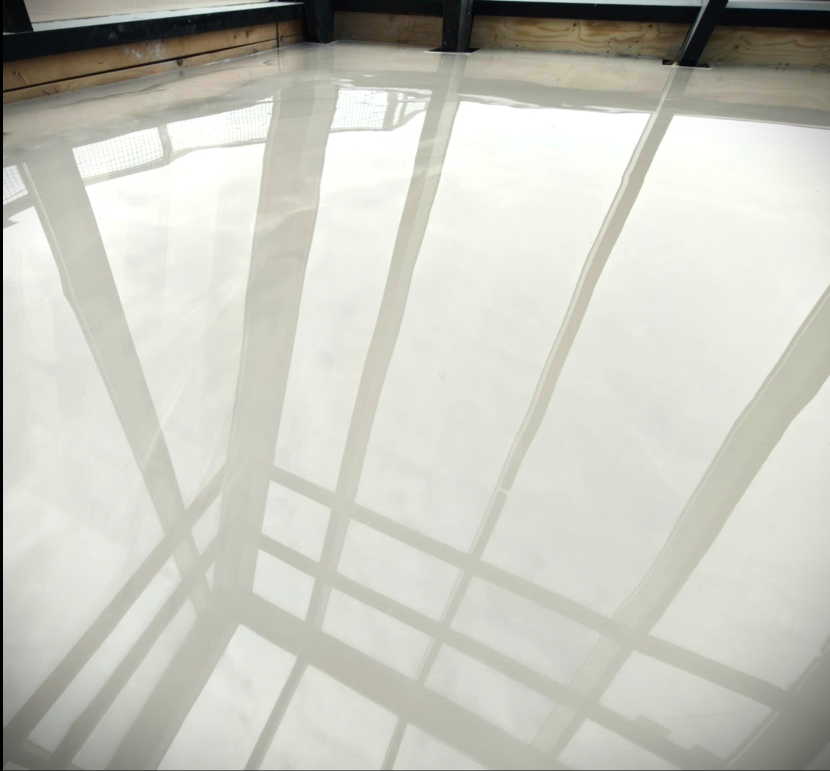 Błyszcząca, jasna posadzka w industrialnym wnętrzu. Widoczne odbicia konstrukcji sufitu, podkreślające gładką powierzchnię. Minimalistyczny design.