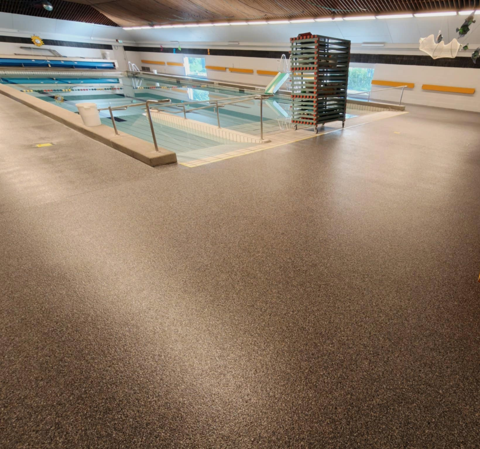 Ciemna, gładka posadzka żywiczna w hali basenowej, widok na basen i metalowy regał na akcesoria. Nowoczesne wykończenie podłogi w obiekcie sportowym.