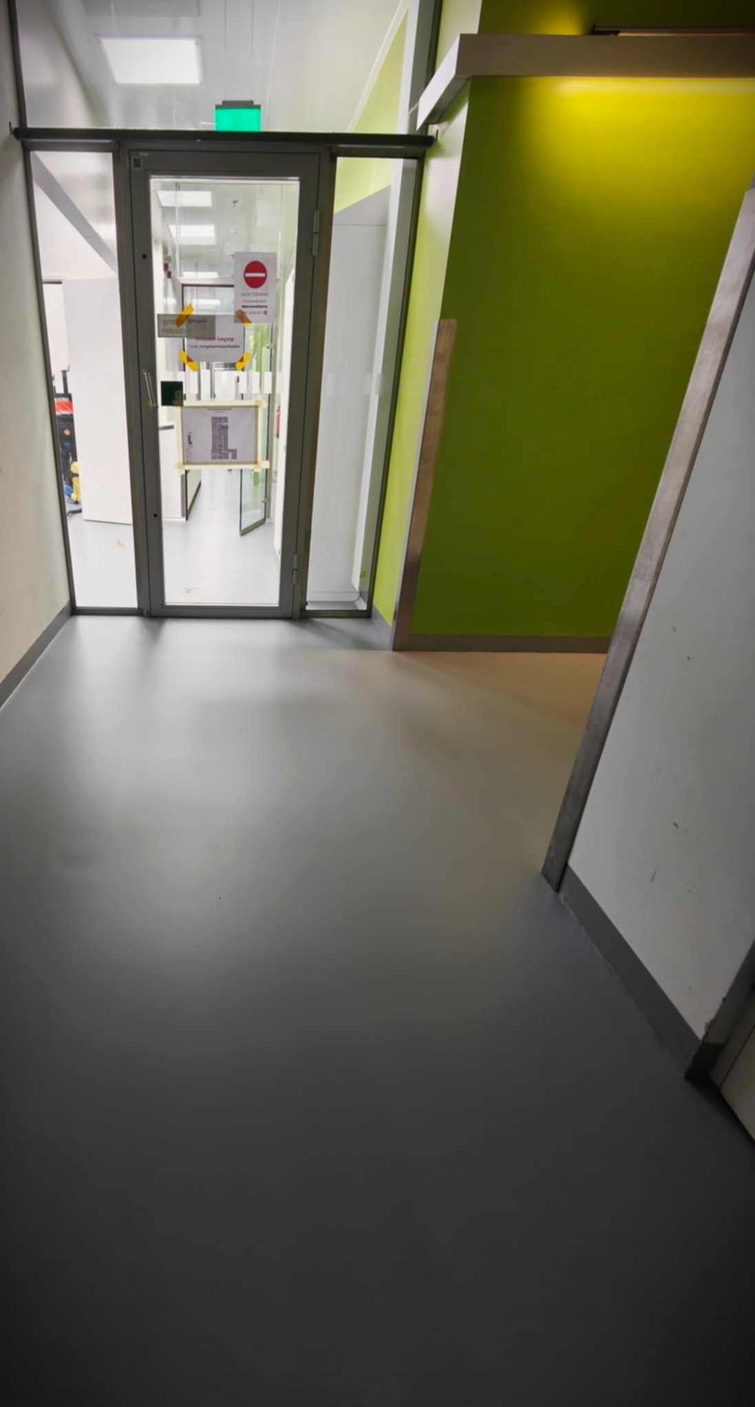 Szara, gładka podłoga w korytarzu z zieloną ścianą w Michałowicach. Widoczne drzwi z oznaczeniami. Minimalistyczny design, jednolite wykończenie.